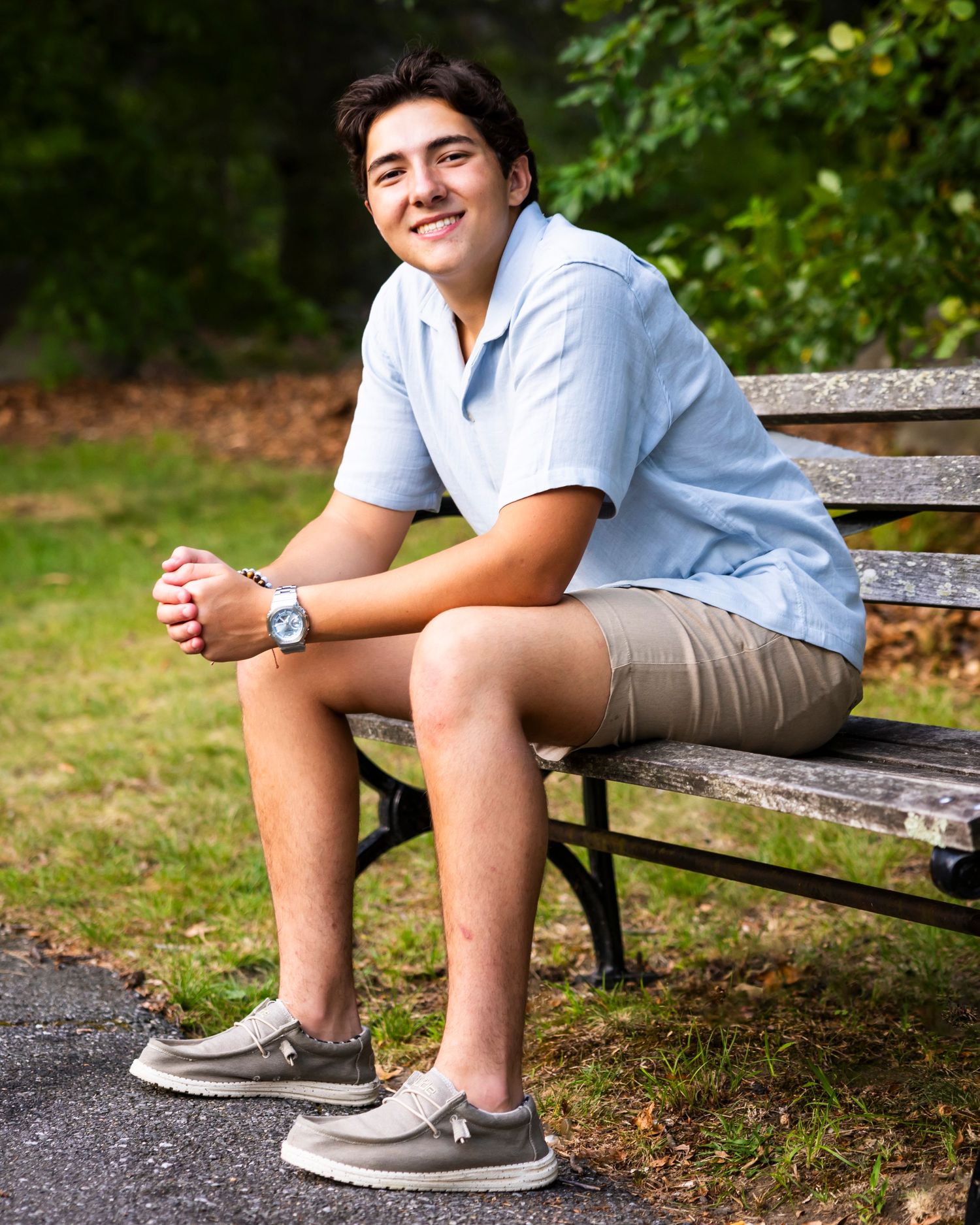 Smiling teenage boy in a light blue polo and khaki shorts sitting on a park bench outdoors.