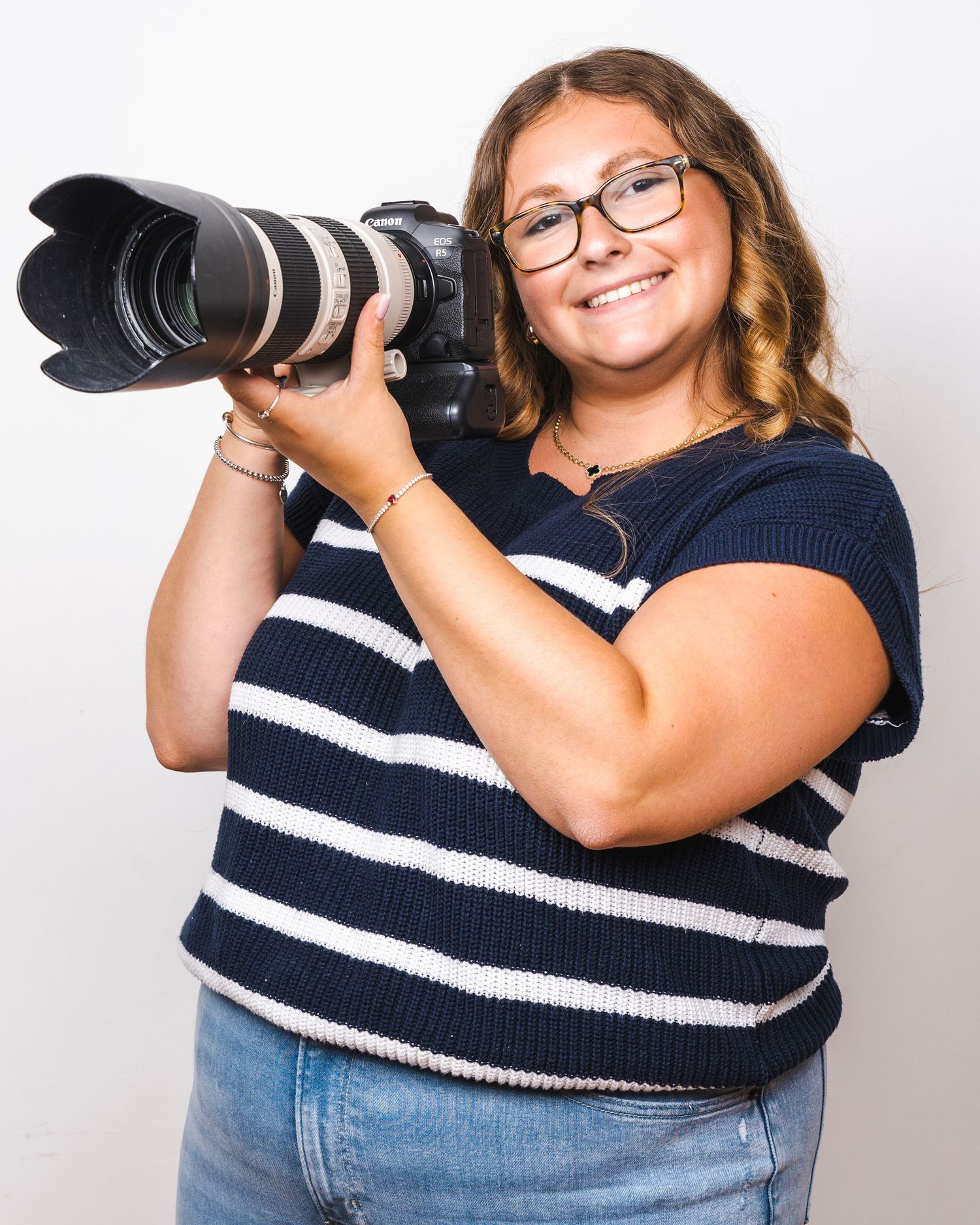 Smiling young woman in striped shirt holding a professional Canon DSLR camera with telephoto lens.