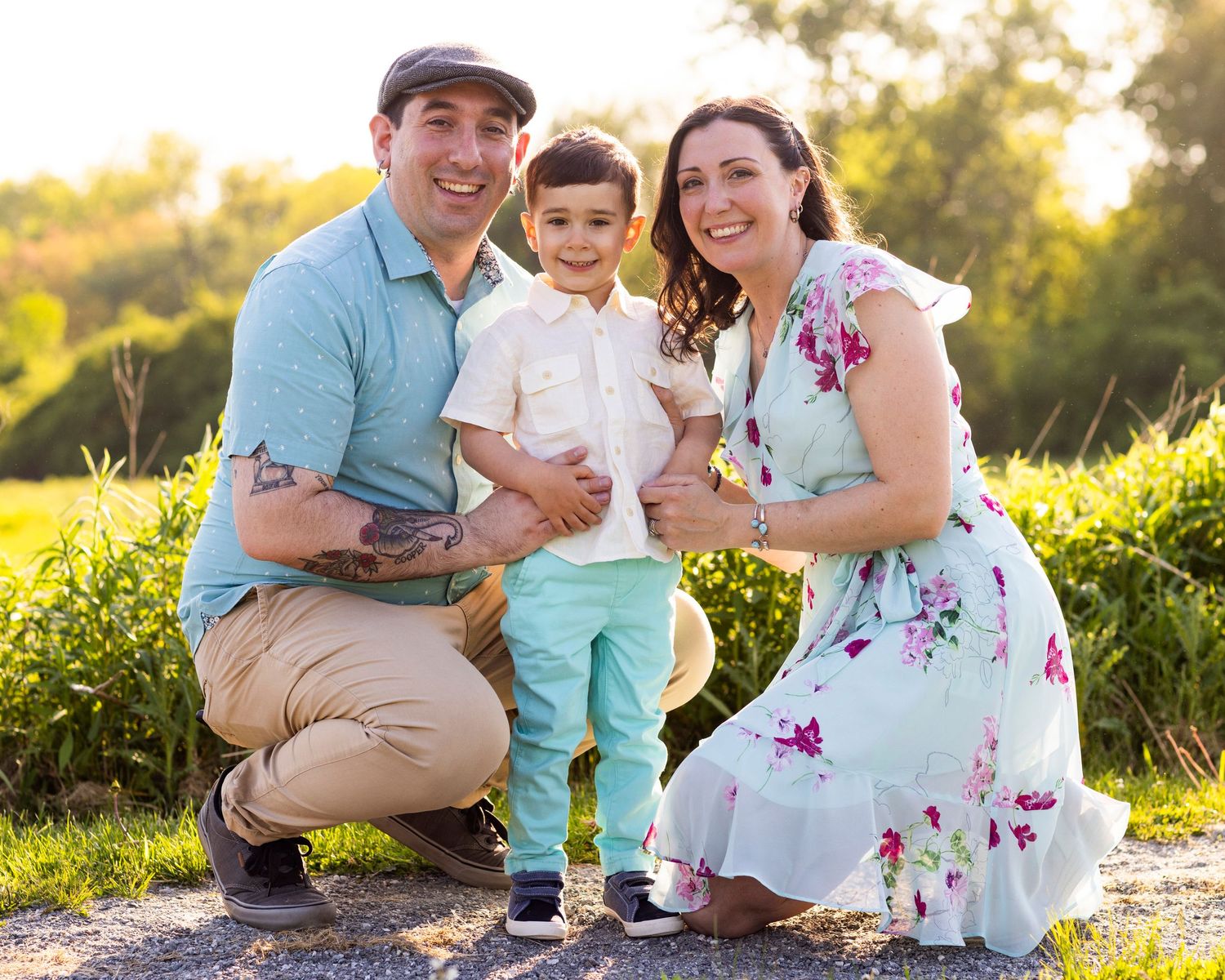 Happy family of three posing outdoors at golden hour, parents crouching beside their young son in green field.