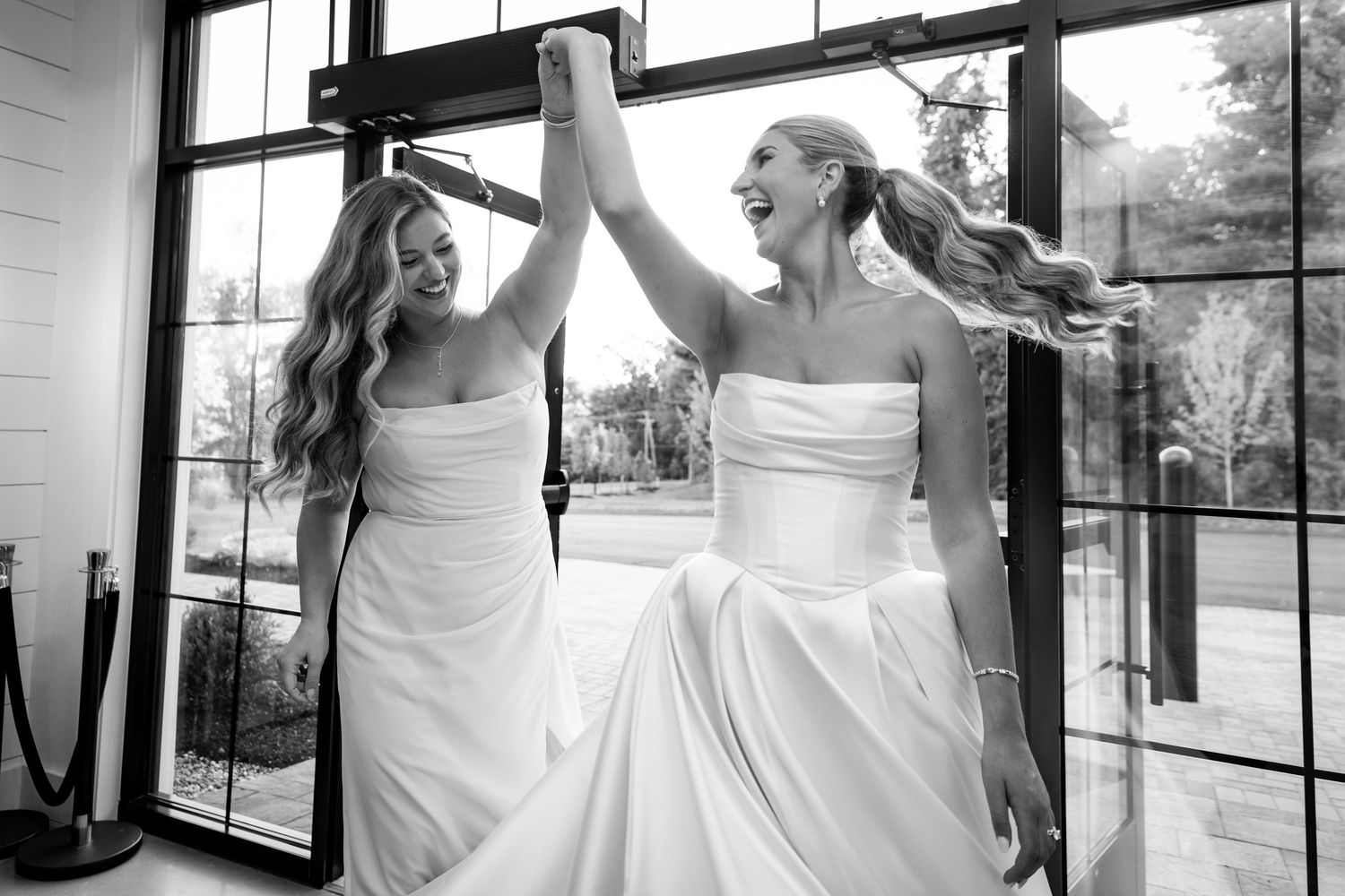 Two brides celebrate joyfully holding hands in black and white wedding portrait.