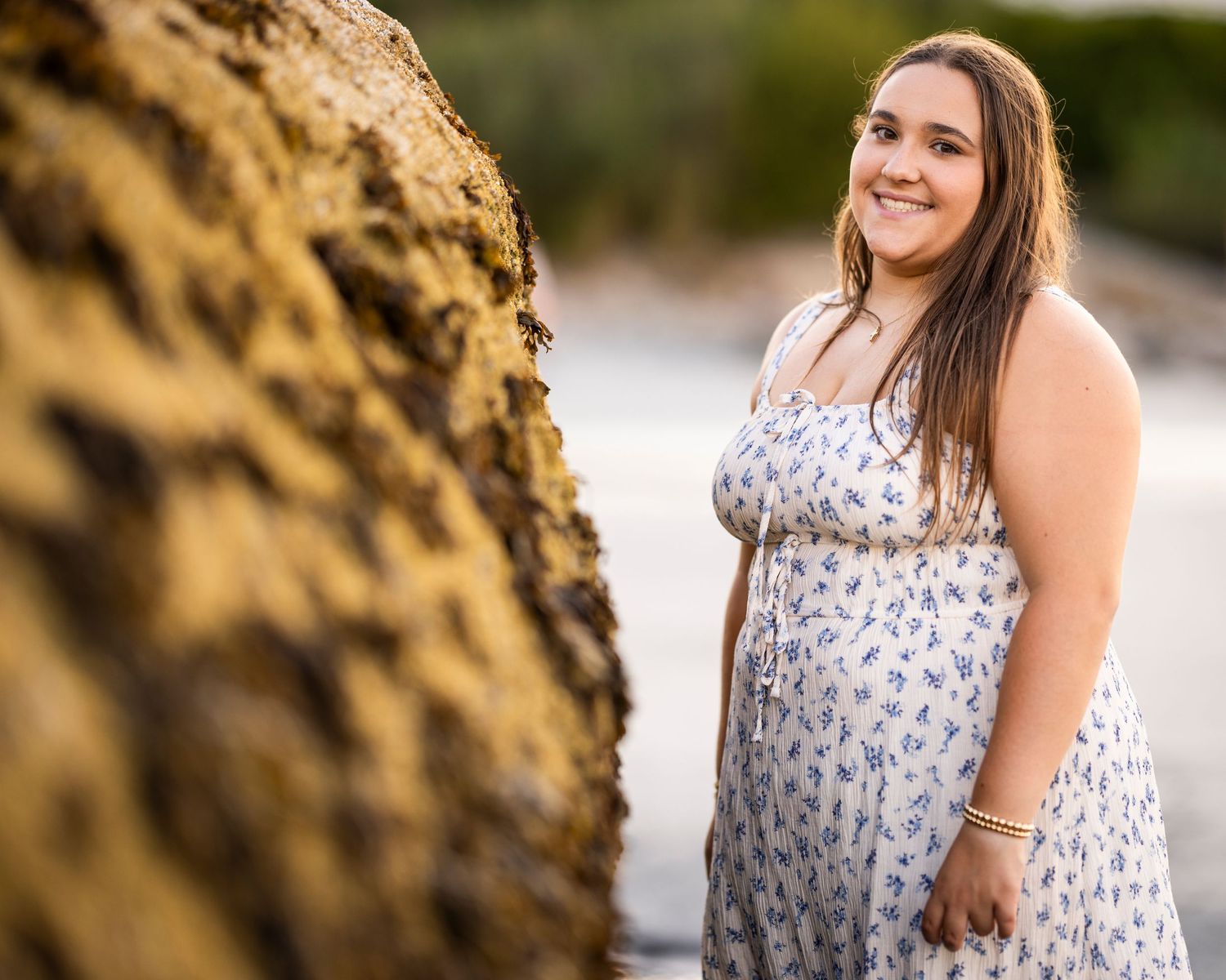 Smiling young woman in a floral sundress posing beside large rocks on a scenic beach at sunset.