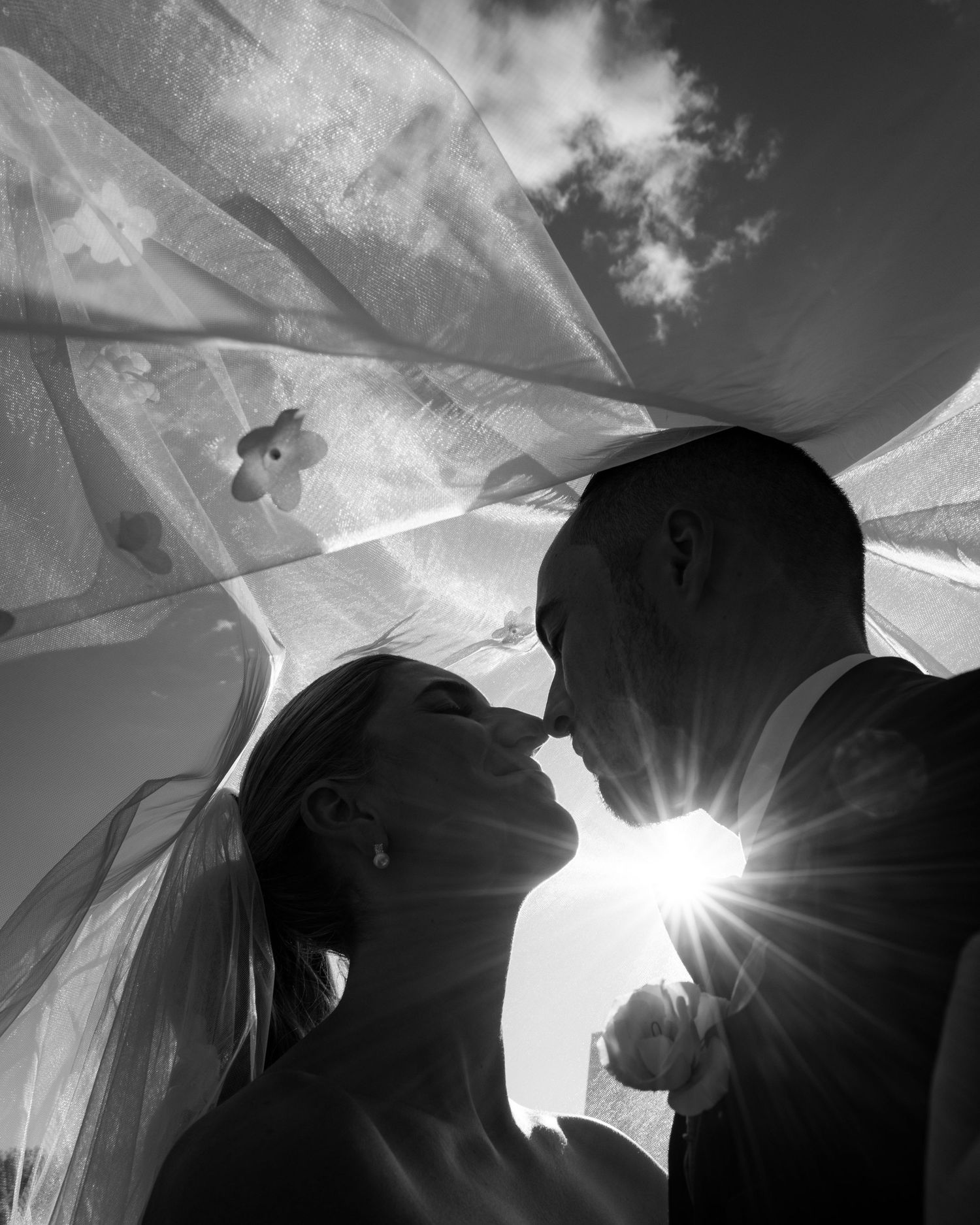 Black and white artistic photo of bride and groom kissing under veil with sunburst and clouds above.