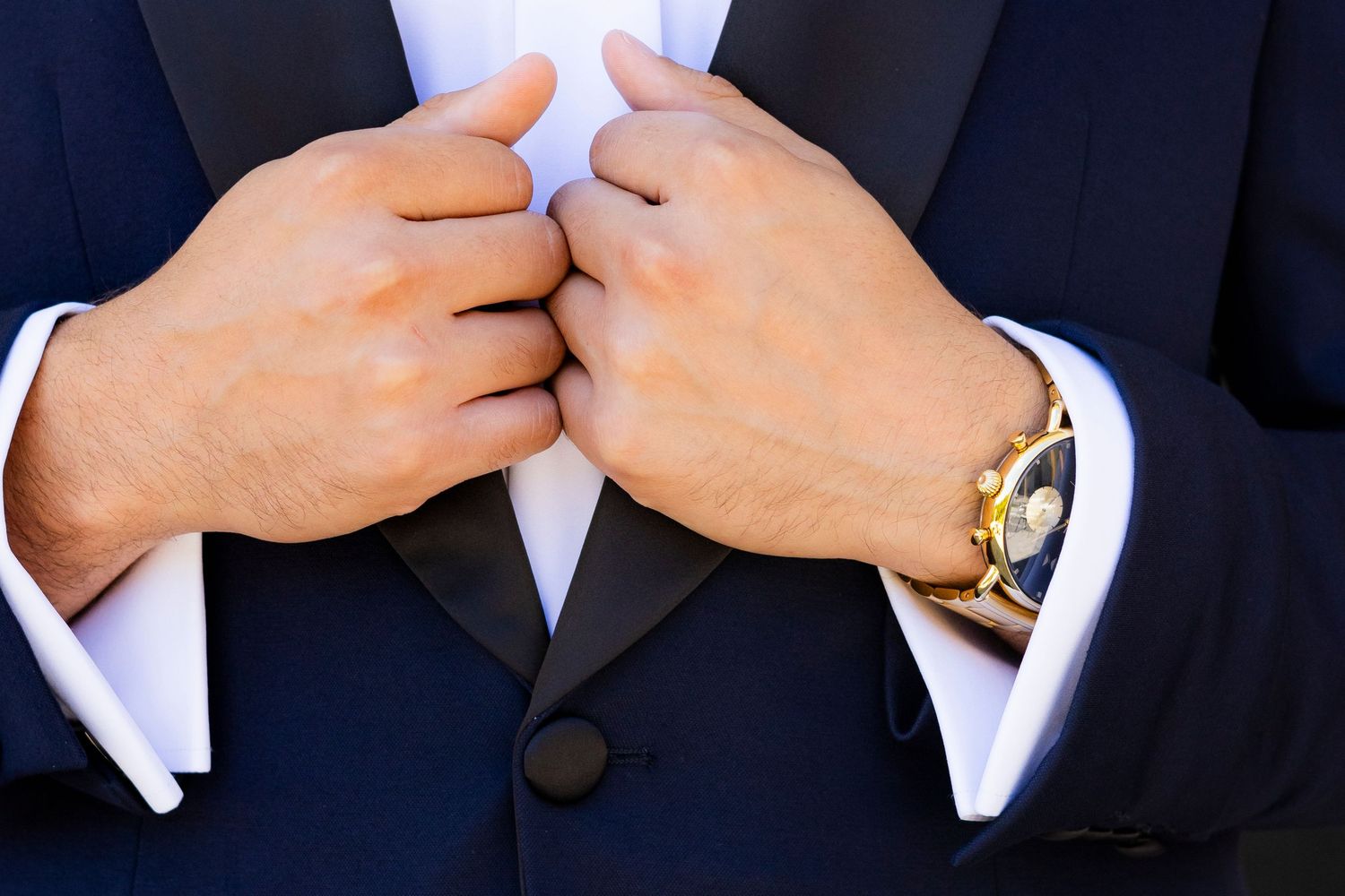 Close-up of groom adjusting navy tuxedo lapels with gold watch on wrist.