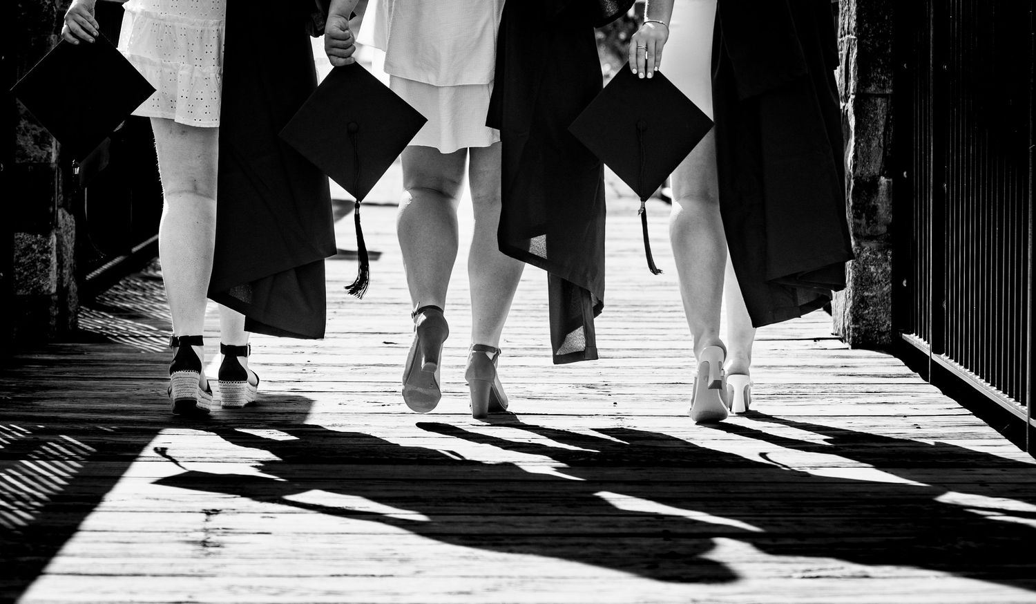 Black and white photo of graduates in heels walking on wooden boardwalk holding mortarboard caps.