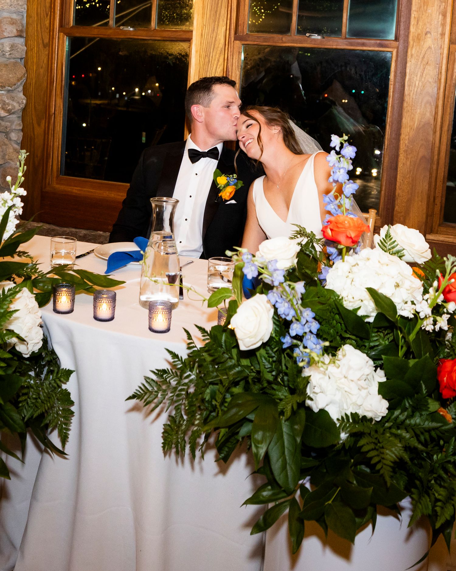 Bride and groom share a sweet kiss at their sweetheart table adorned with lush white floral arrangements.