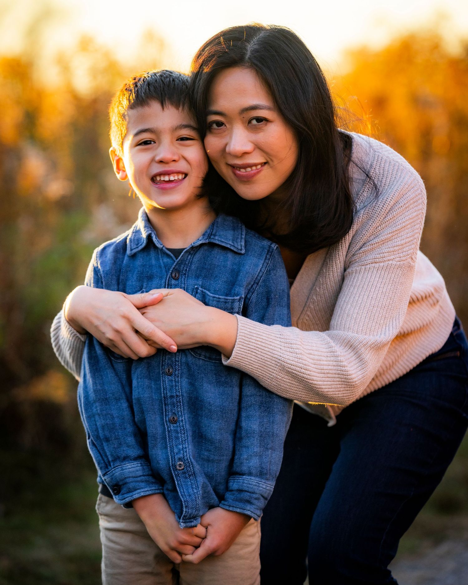 A mother hugs her smiling young son outdoors during a warm golden hour family portrait session.