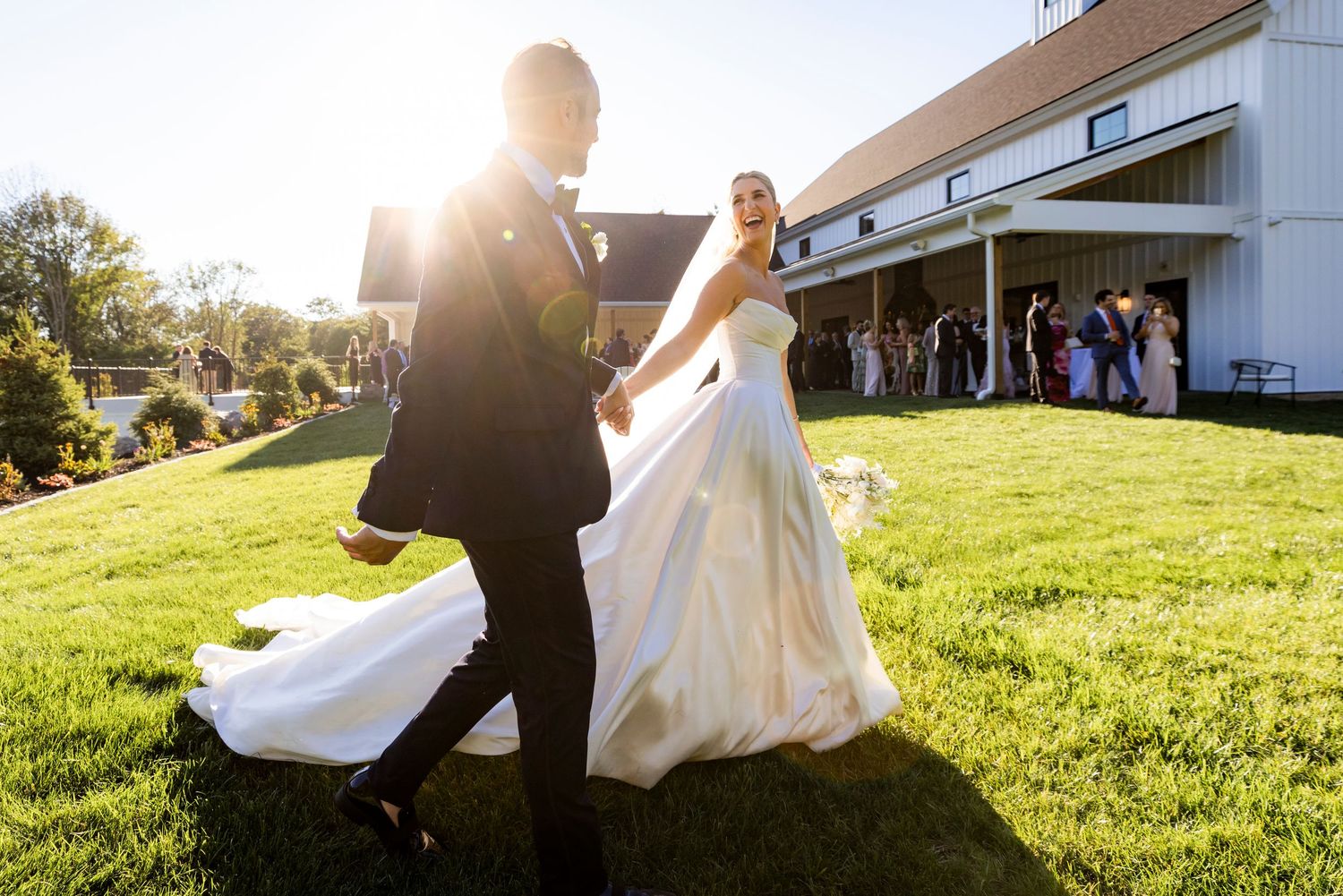 Bride and groom walk hand in hand across green lawn toward wedding venue guests.