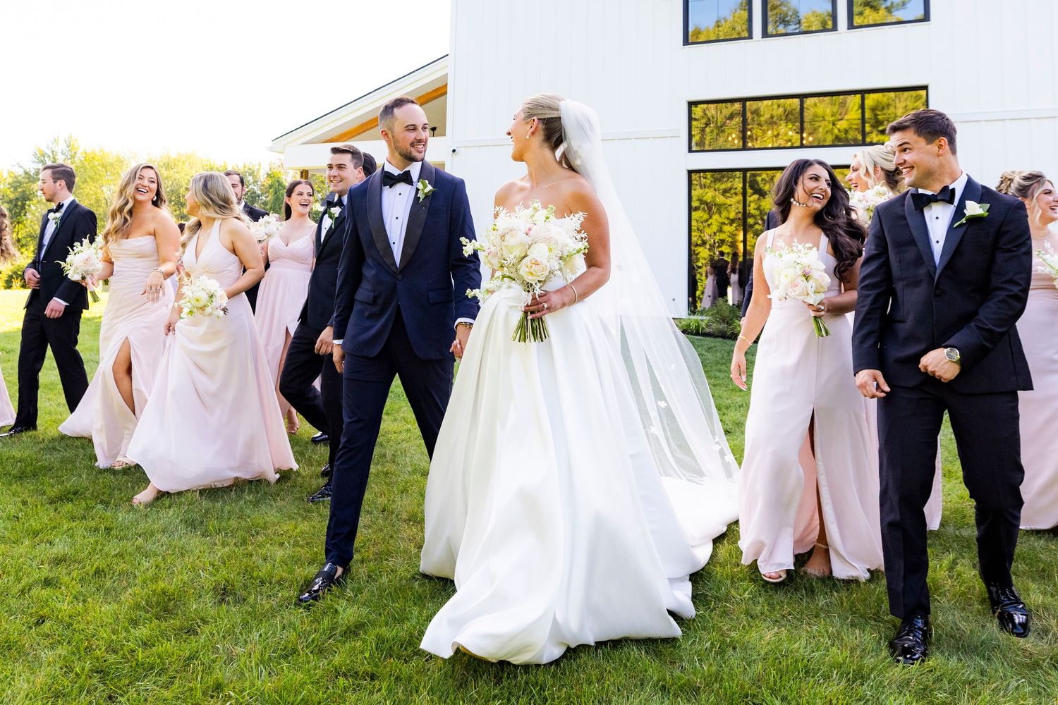 Bride and groom leading wedding party across green lawn with bridesmaids in pink dresses and groomsmen in tuxedos.