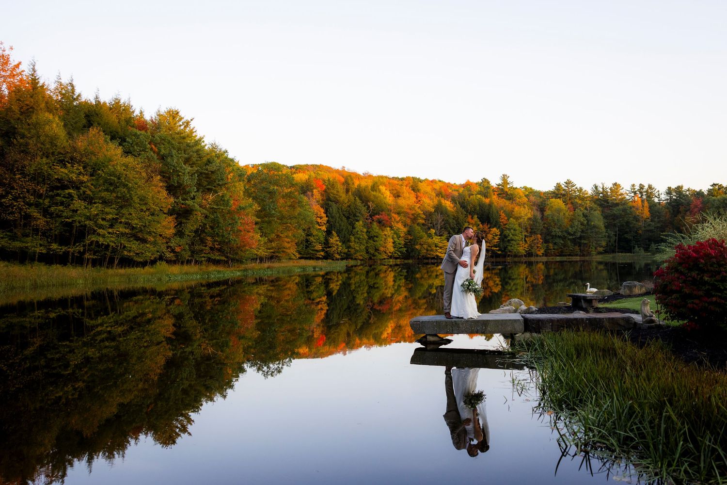 A couple embraces on a lakeside dock surrounded by stunning fall foliage reflected perfectly in the still water.