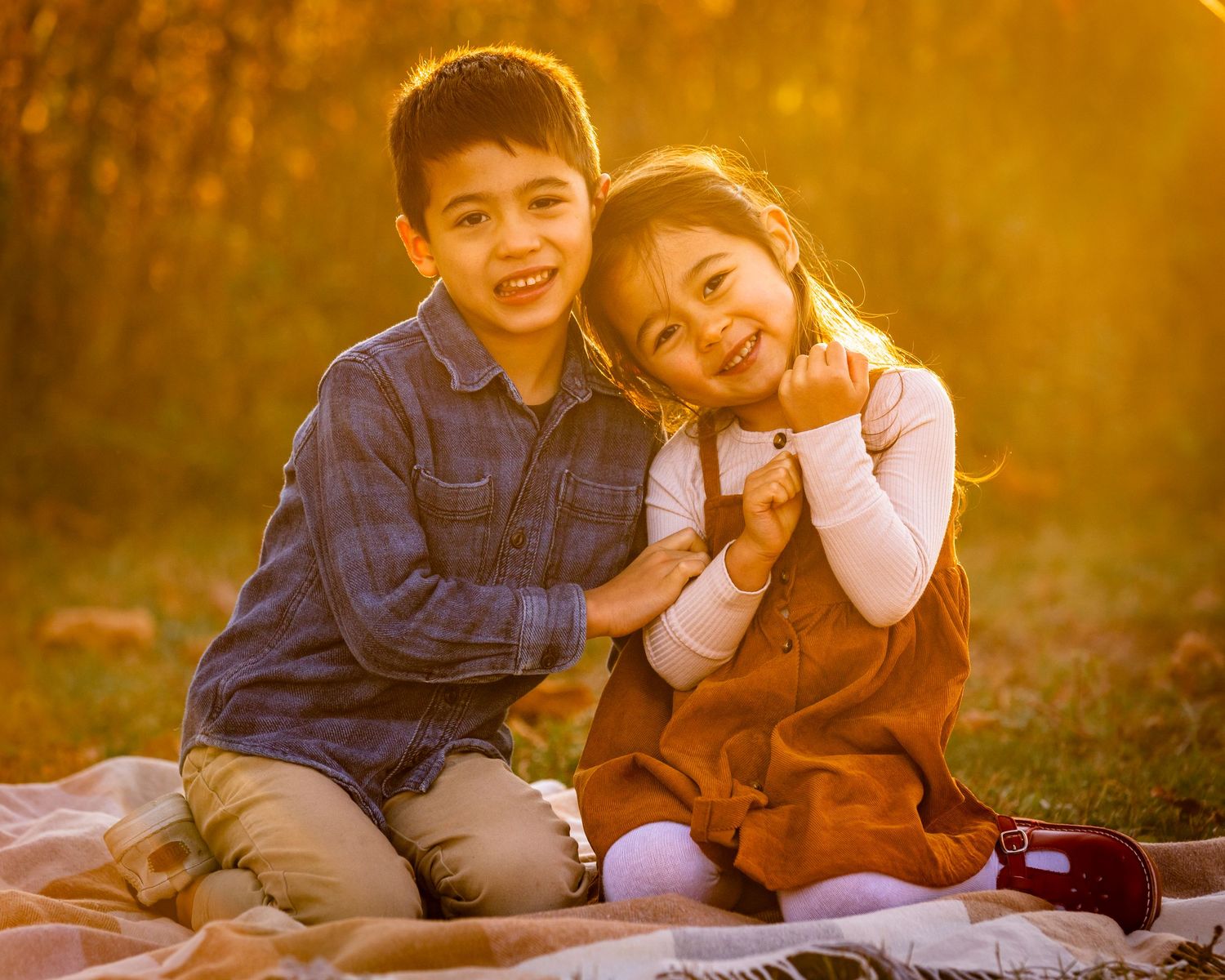 Two young siblings smile and hug on a blanket outdoors during a golden autumn sunset photo session.