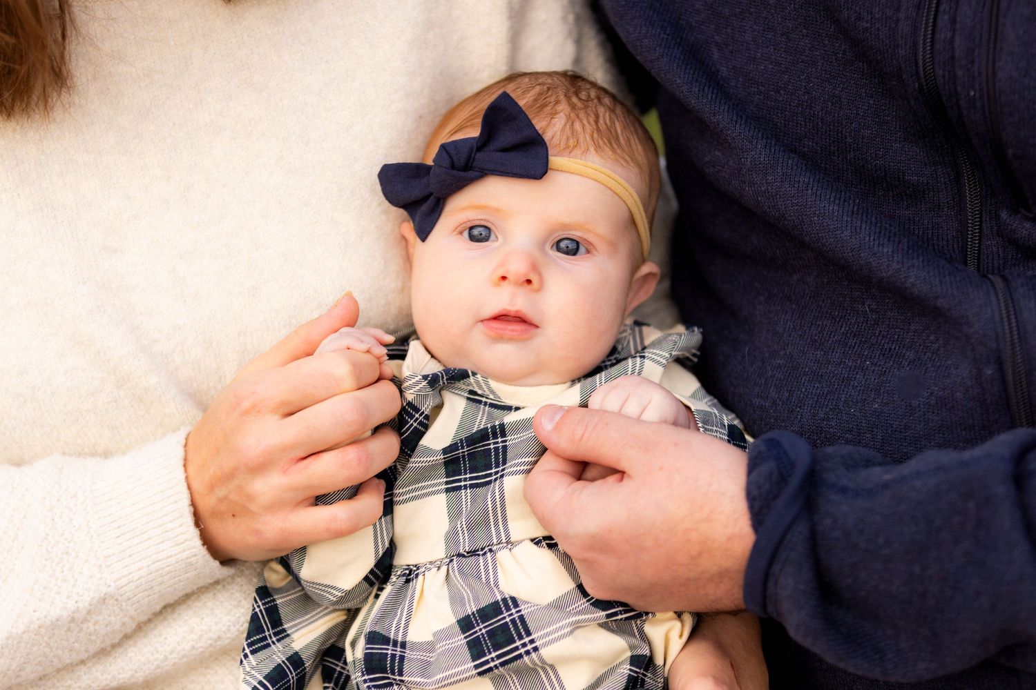 Adorable baby girl in plaid dress with navy bow headband held by parents during newborn session.