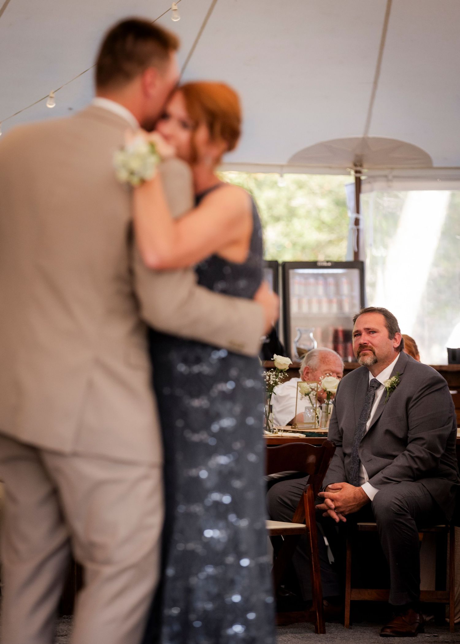 Groom shares a dance with mother while a seated guest watches during wedding reception under tent.