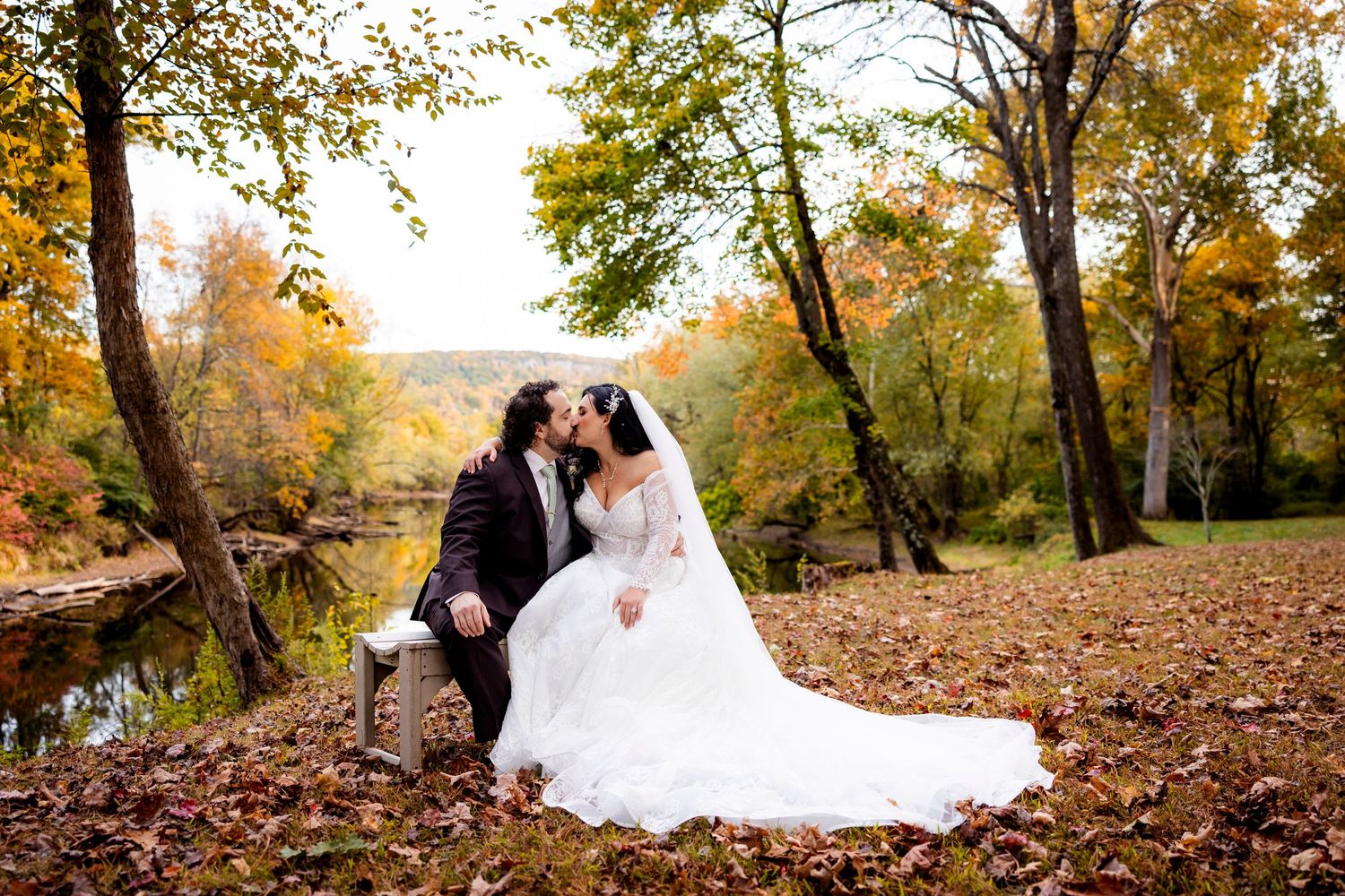 Bride and groom sharing a kiss among fallen autumn leaves beside a serene river in a colorful forest.