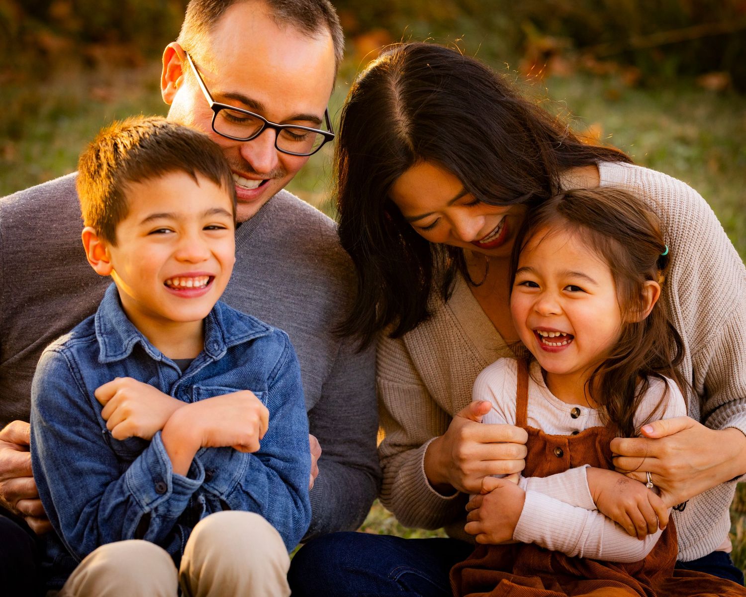 A joyful family of four laughs together while sitting outdoors during a warm autumn photo session.