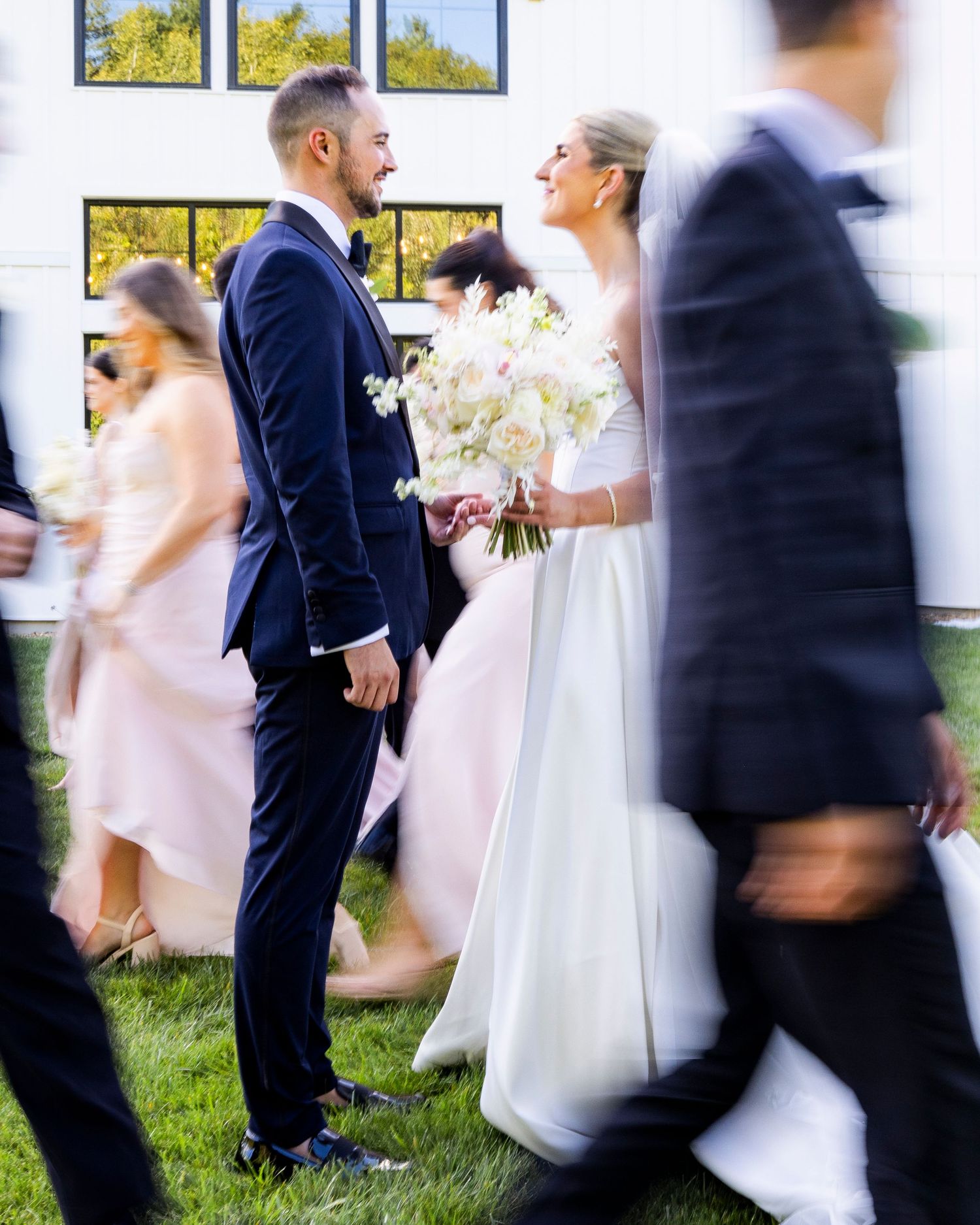 Bride holding white bouquet gazes at groom in navy suit with blurred wedding guests walking past them.