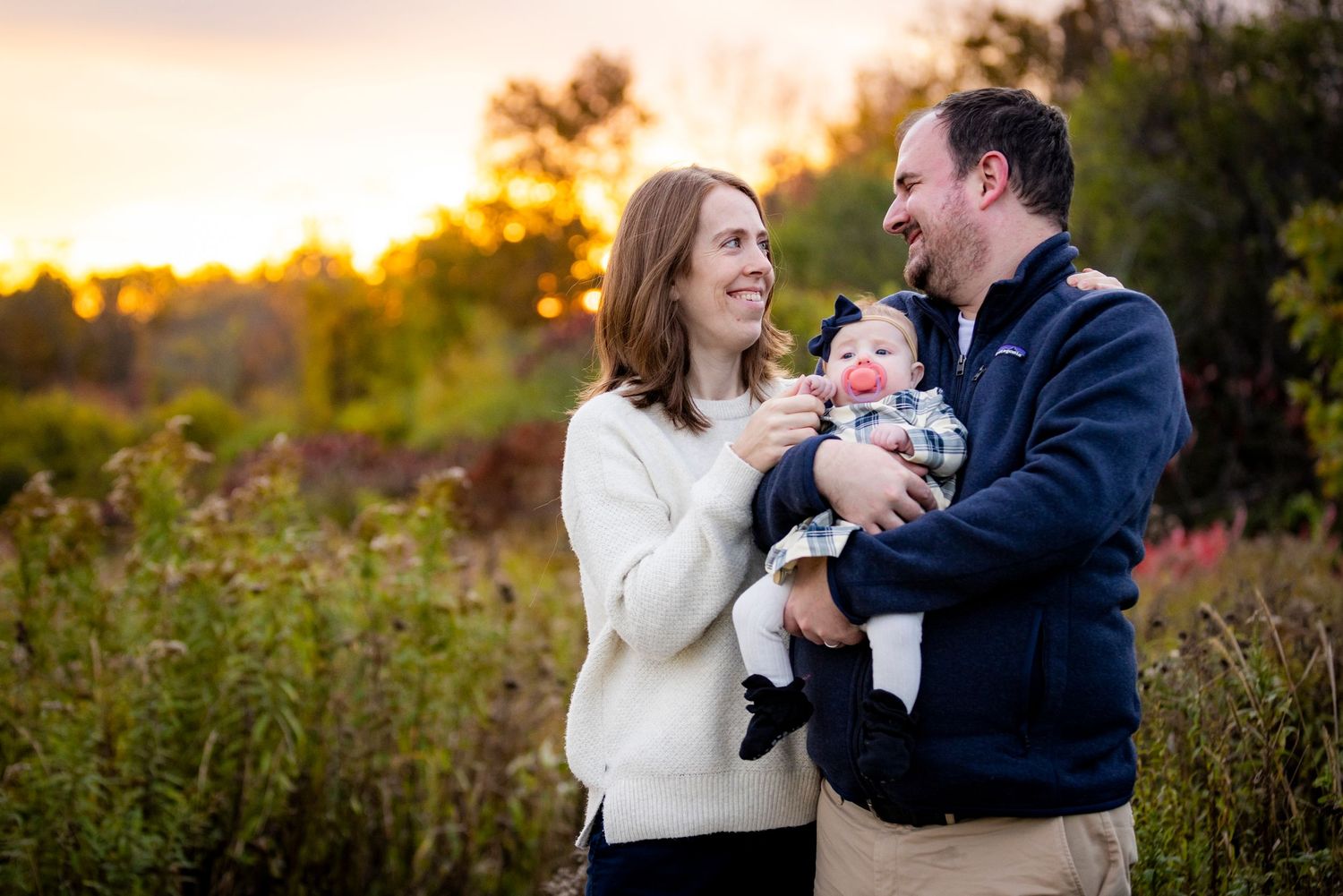 Joyful parents smile and laugh with their happy baby during a colorful fall outdoor photo session.