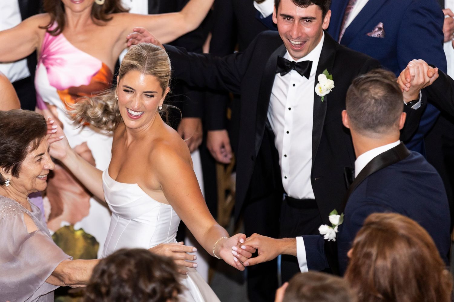 Bride and groom hold hands dancing joyfully surrounded by celebrating guests at wedding reception.