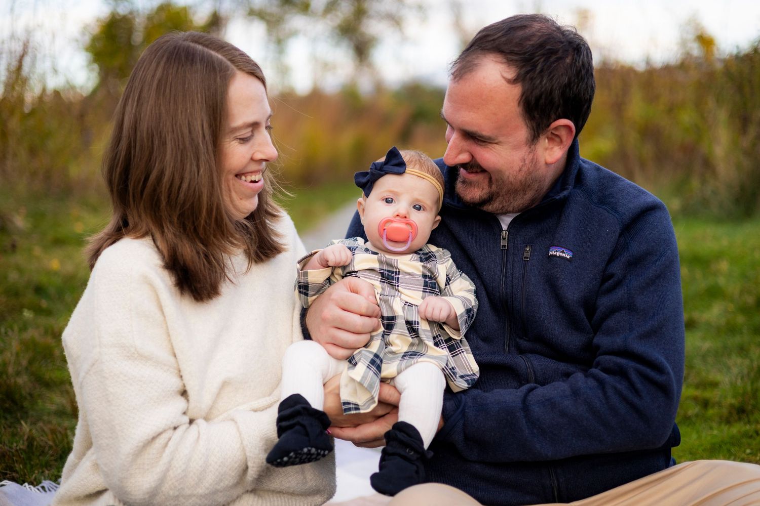 Young couple smiles lovingly at their baby girl with a pink pacifier during outdoor family photos.