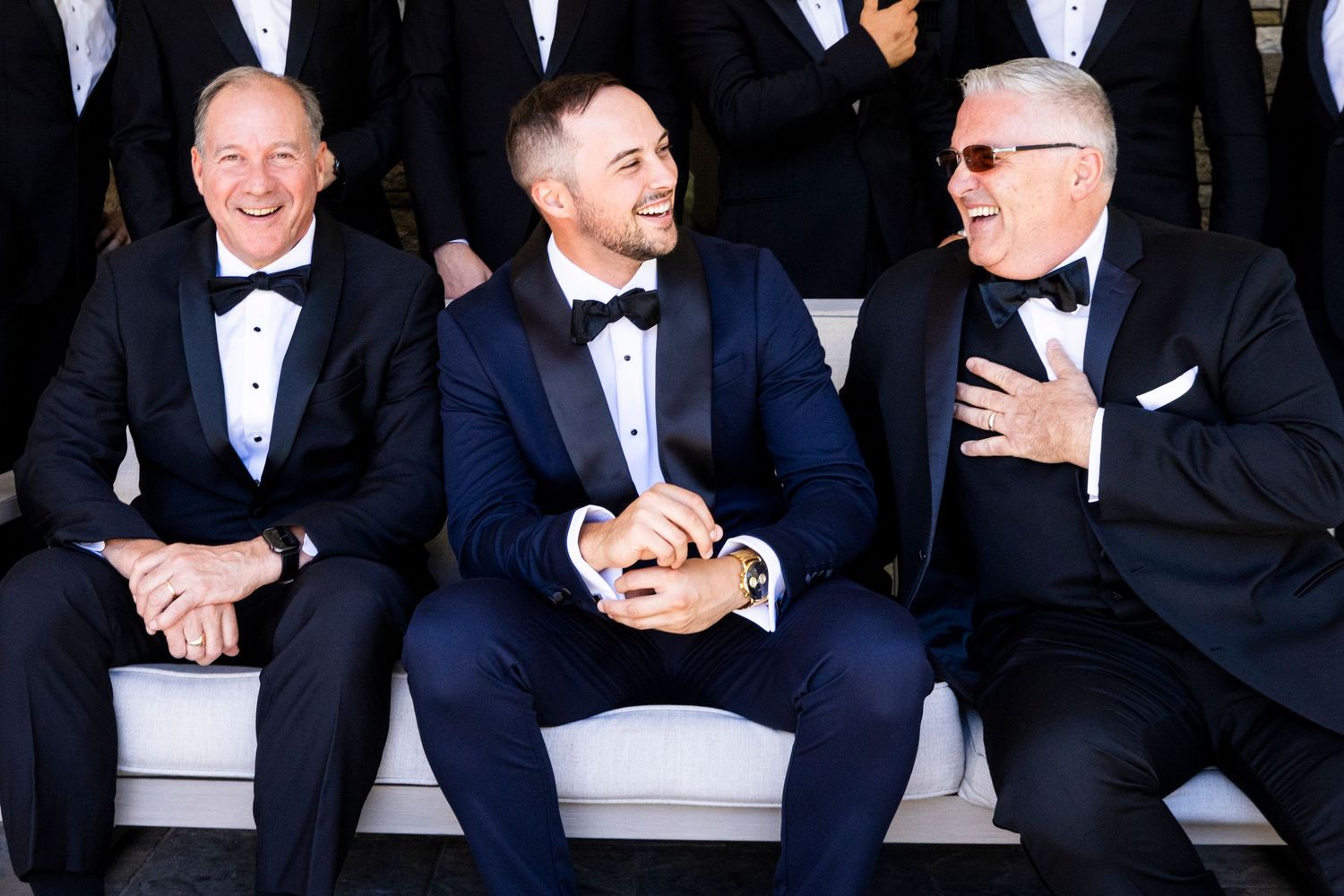 Three men in tuxedos laughing together at wedding reception seated on white bench.