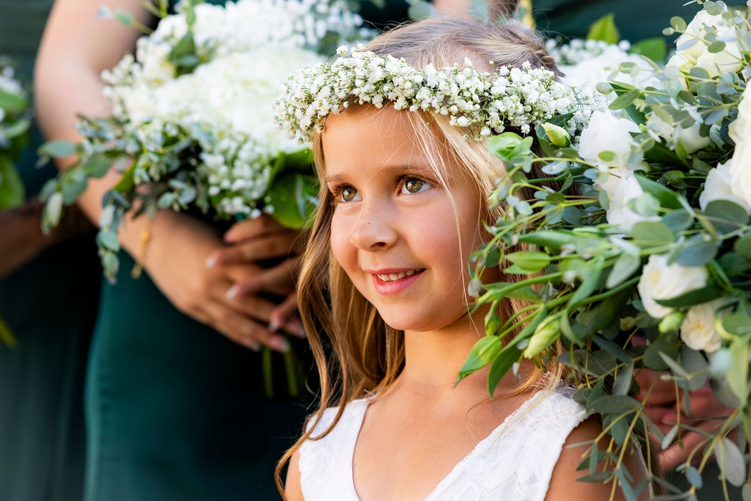 Adorable flower girl wearing white floral crown smiling surrounded by lush wedding bouquets.
