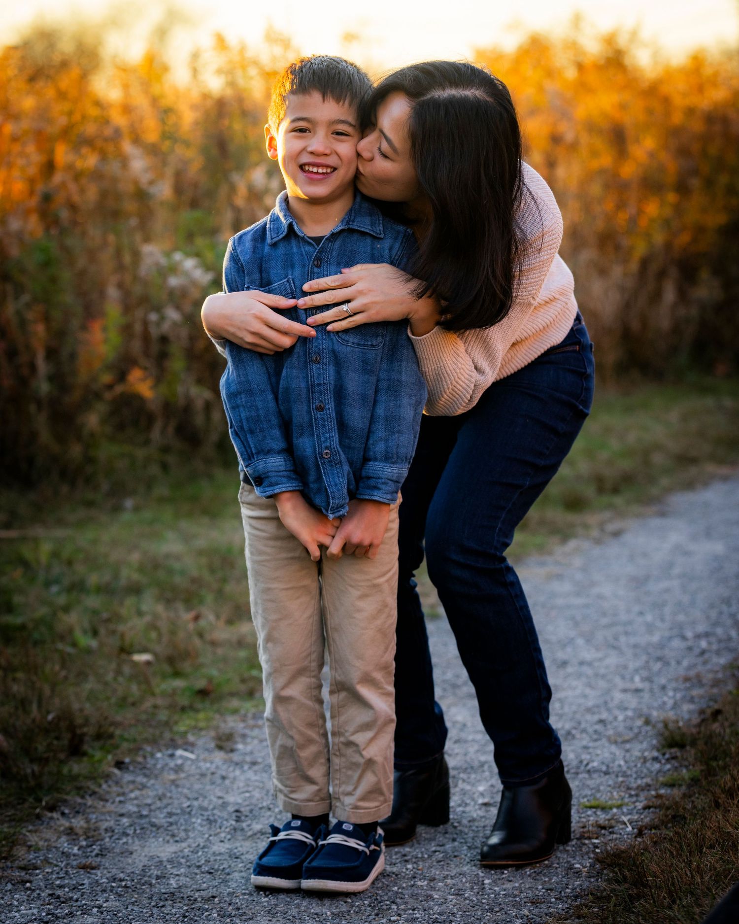 A loving mother kisses her laughing young son on a gravel path surrounded by fall foliage.