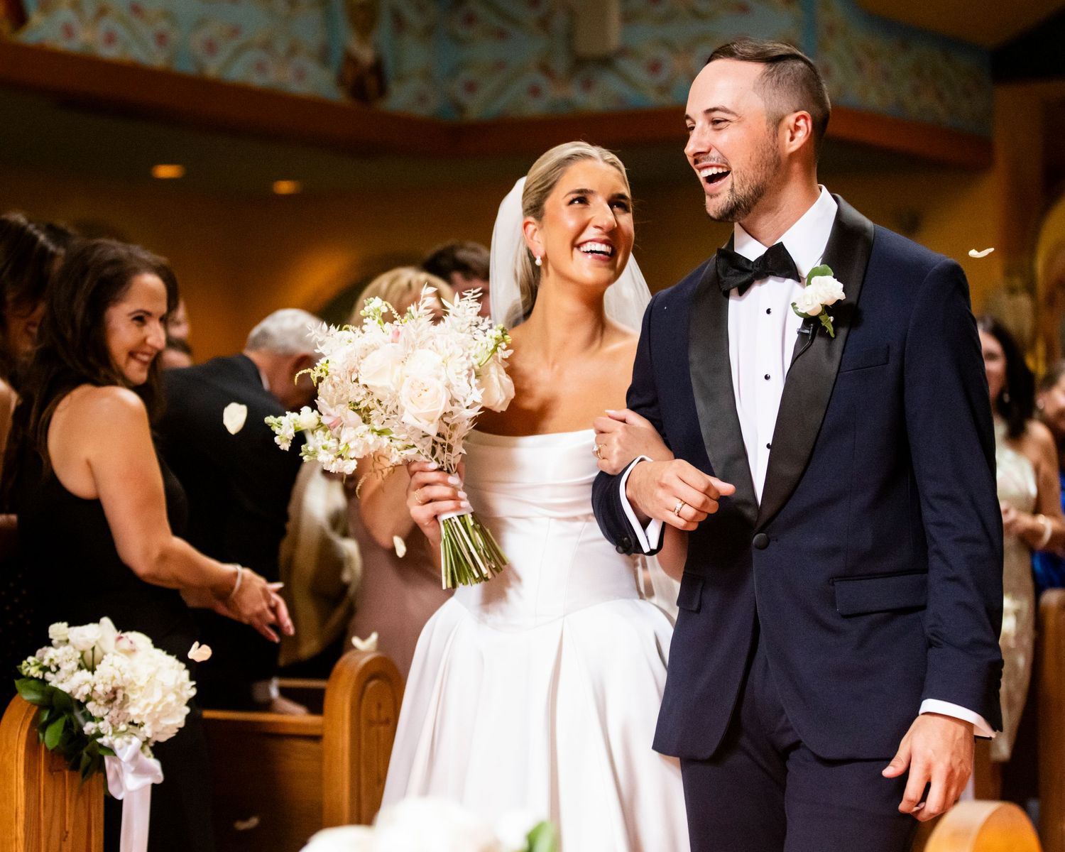 Smiling bride and groom walk down church aisle after wedding ceremony, bride holding white floral bouquet.