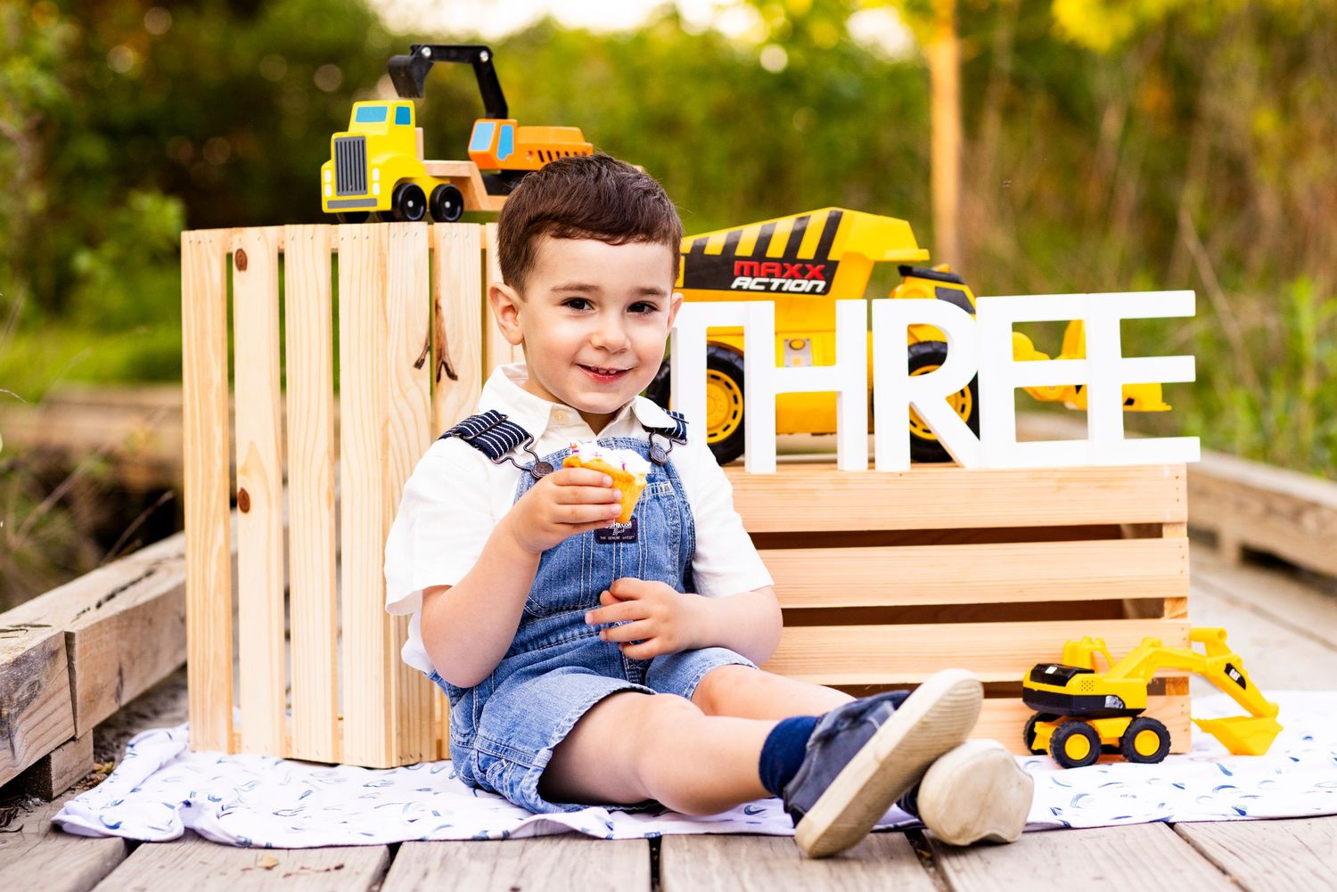 Smiling toddler boy at construction-themed third birthday photo session surrounded by toy trucks and wooden crates.