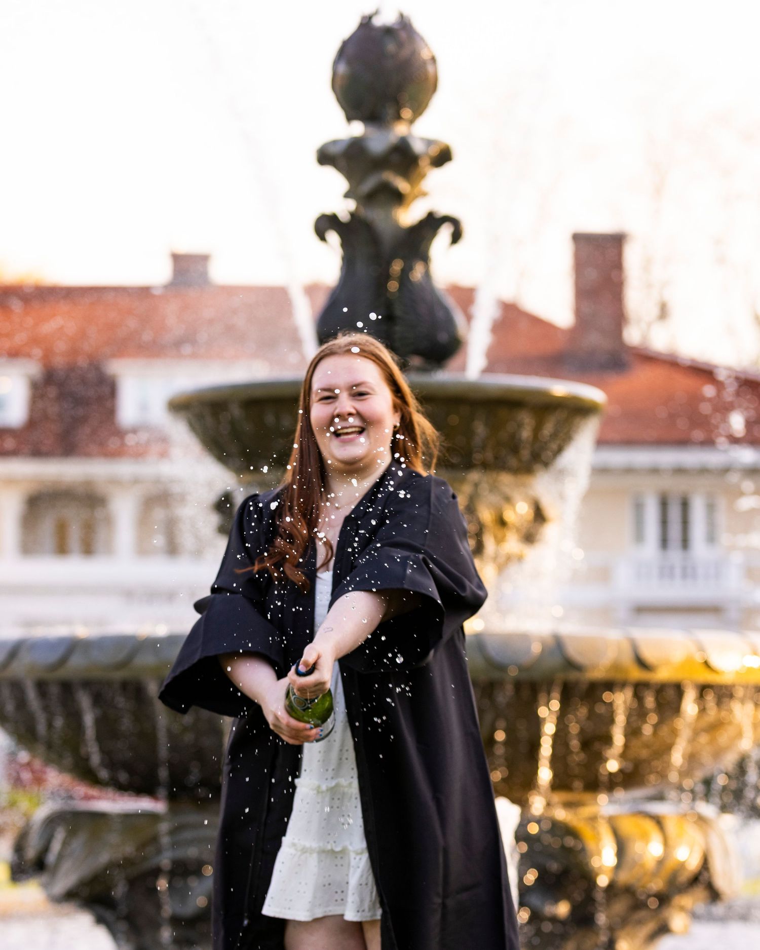 A smiling graduate in a black gown pops a champagne bottle in front of a decorative outdoor fountain.