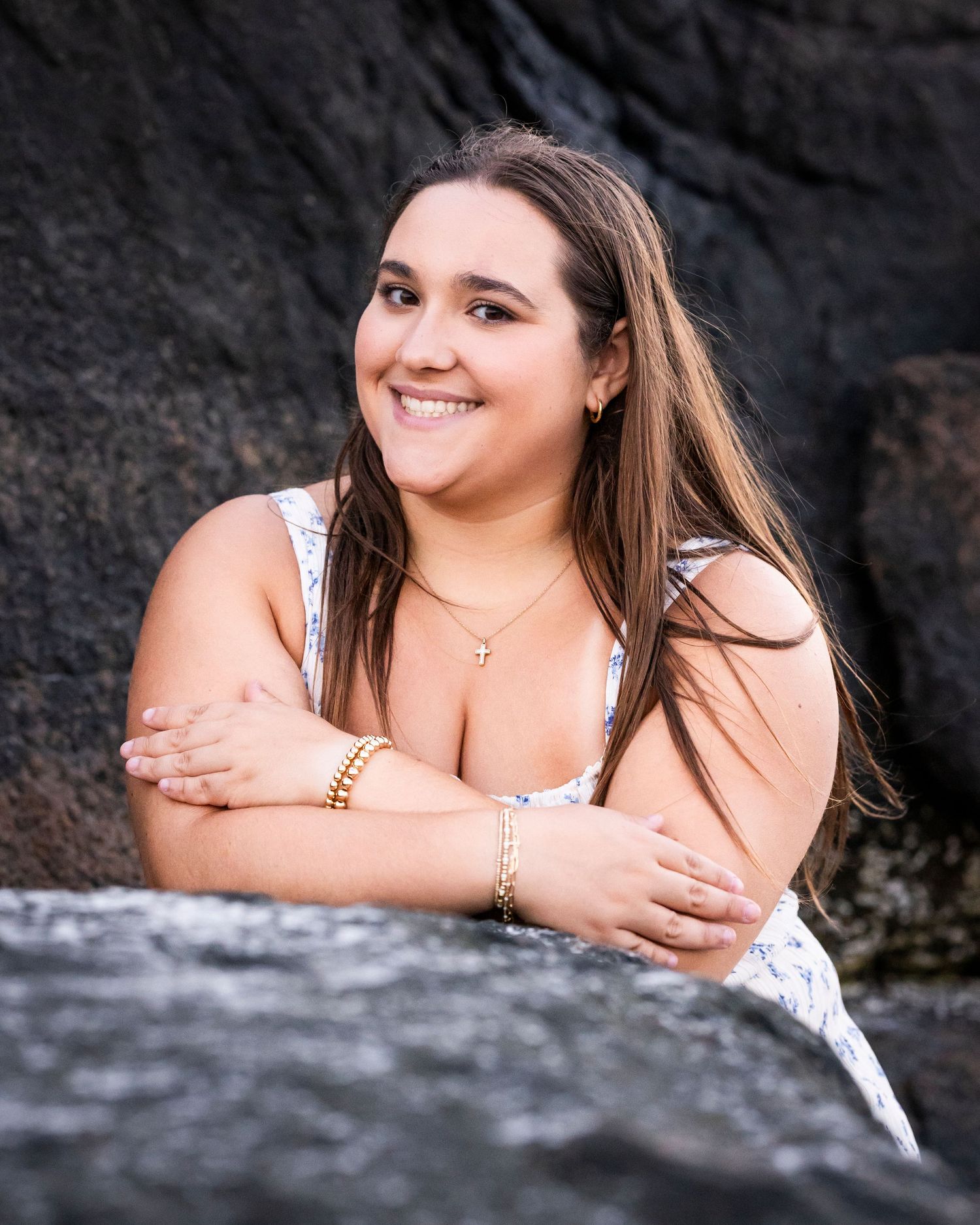 Smiling young woman in white floral dress rests arms on rocky surface in beach portrait.