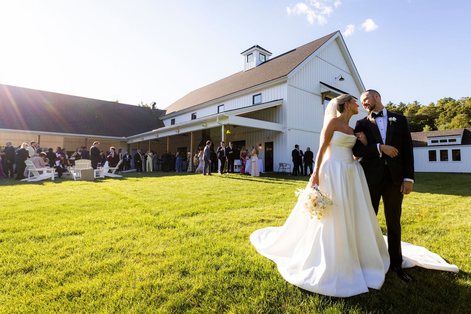 Bride and groom standing on green lawn with white barn wedding venue and guests gathered in background.