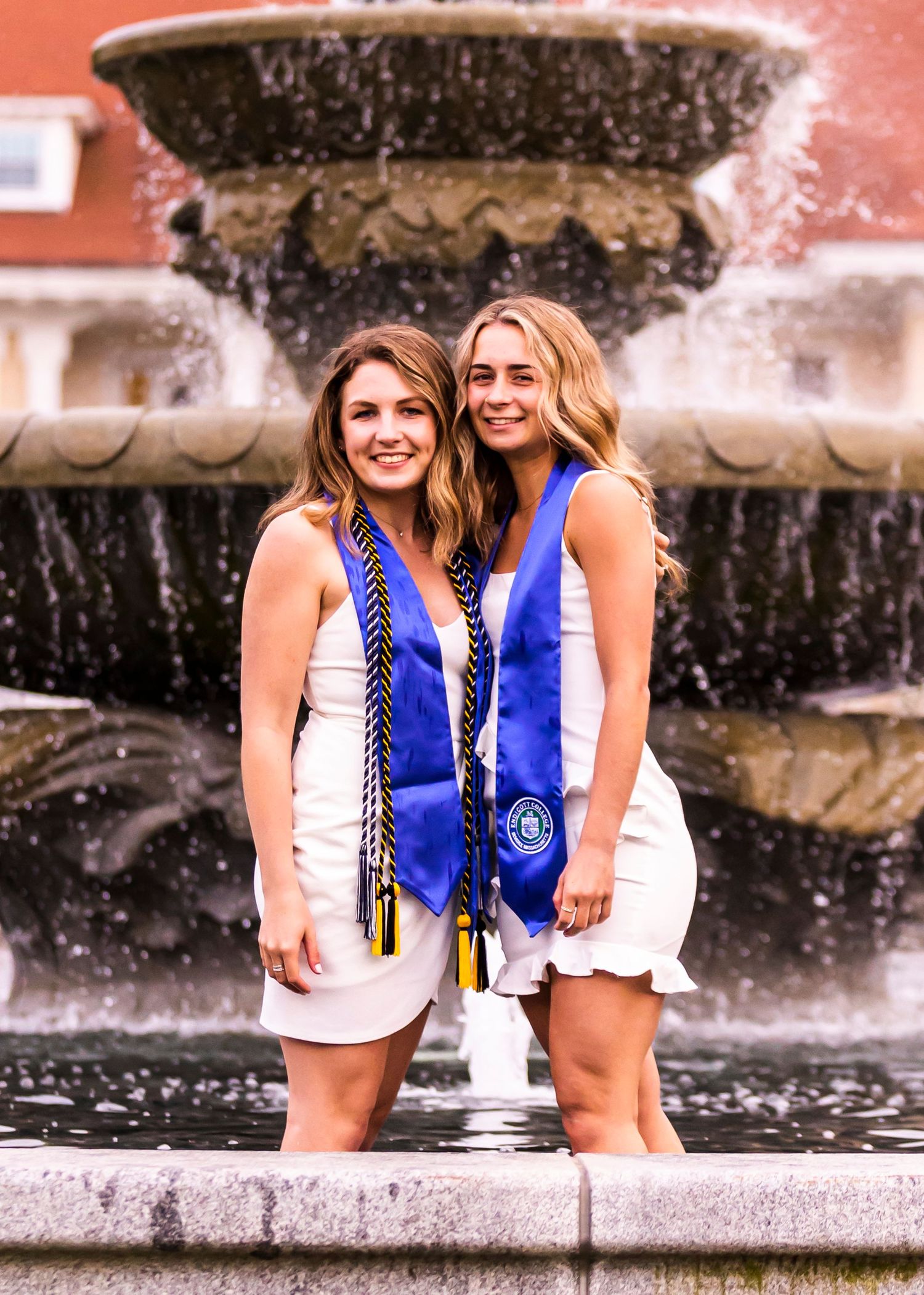 Two smiling female graduates in white dresses wearing blue graduation stoles posing by a decorative fountain.