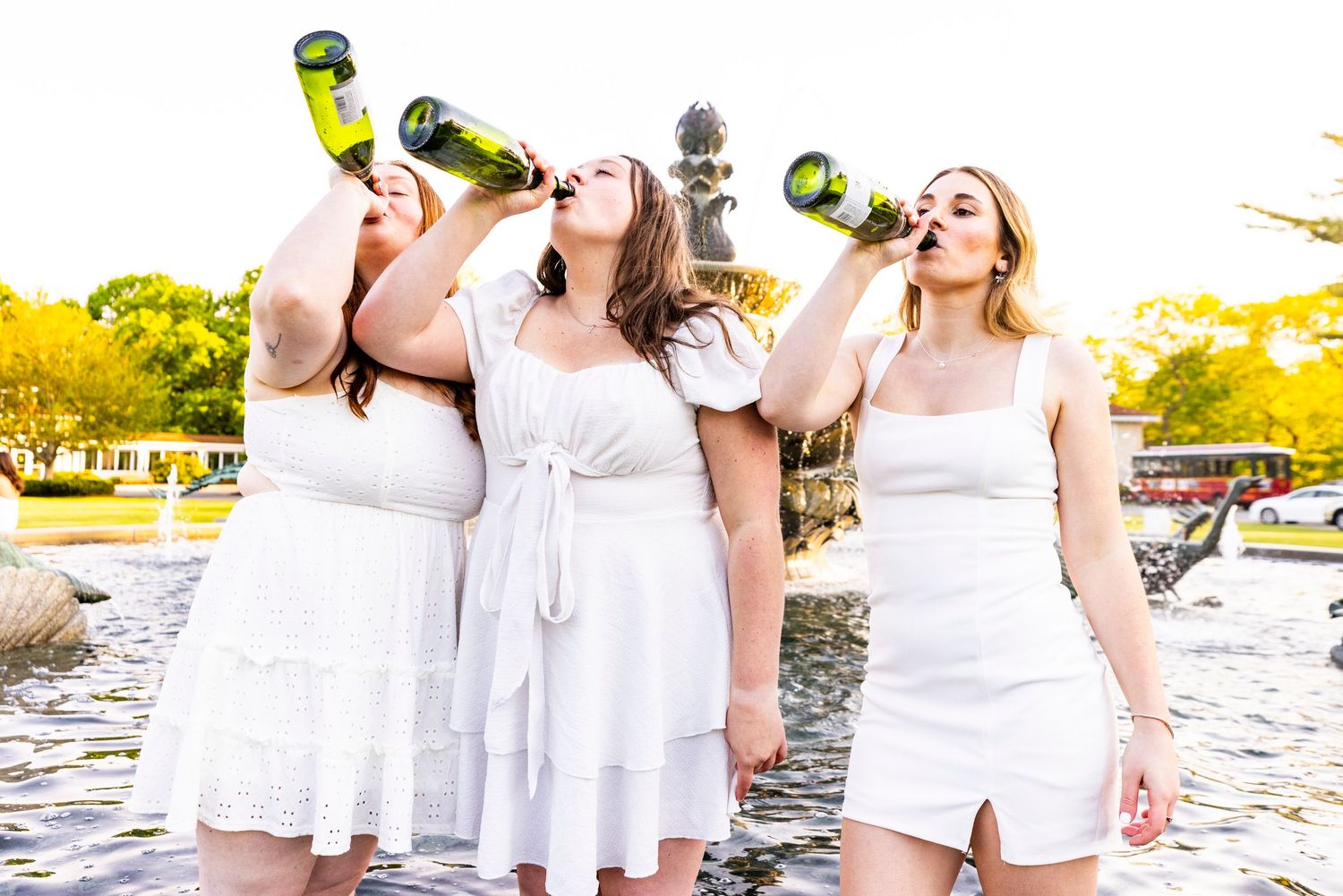 Three young women in white dresses drinking from champagne bottles by campus fountain at graduation.
