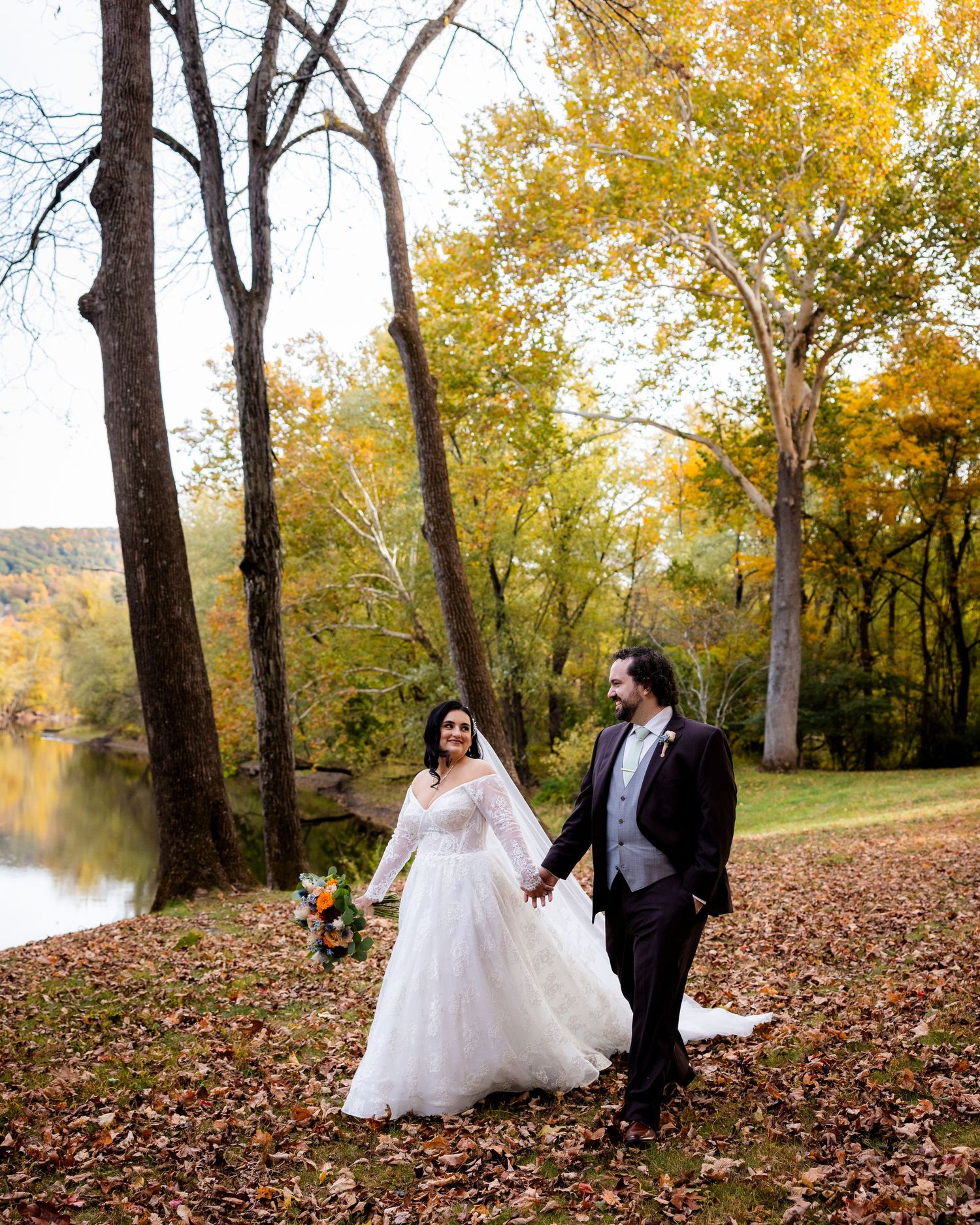 Bride and groom holding hands walking through fallen leaves with golden autumn trees and river backdrop.