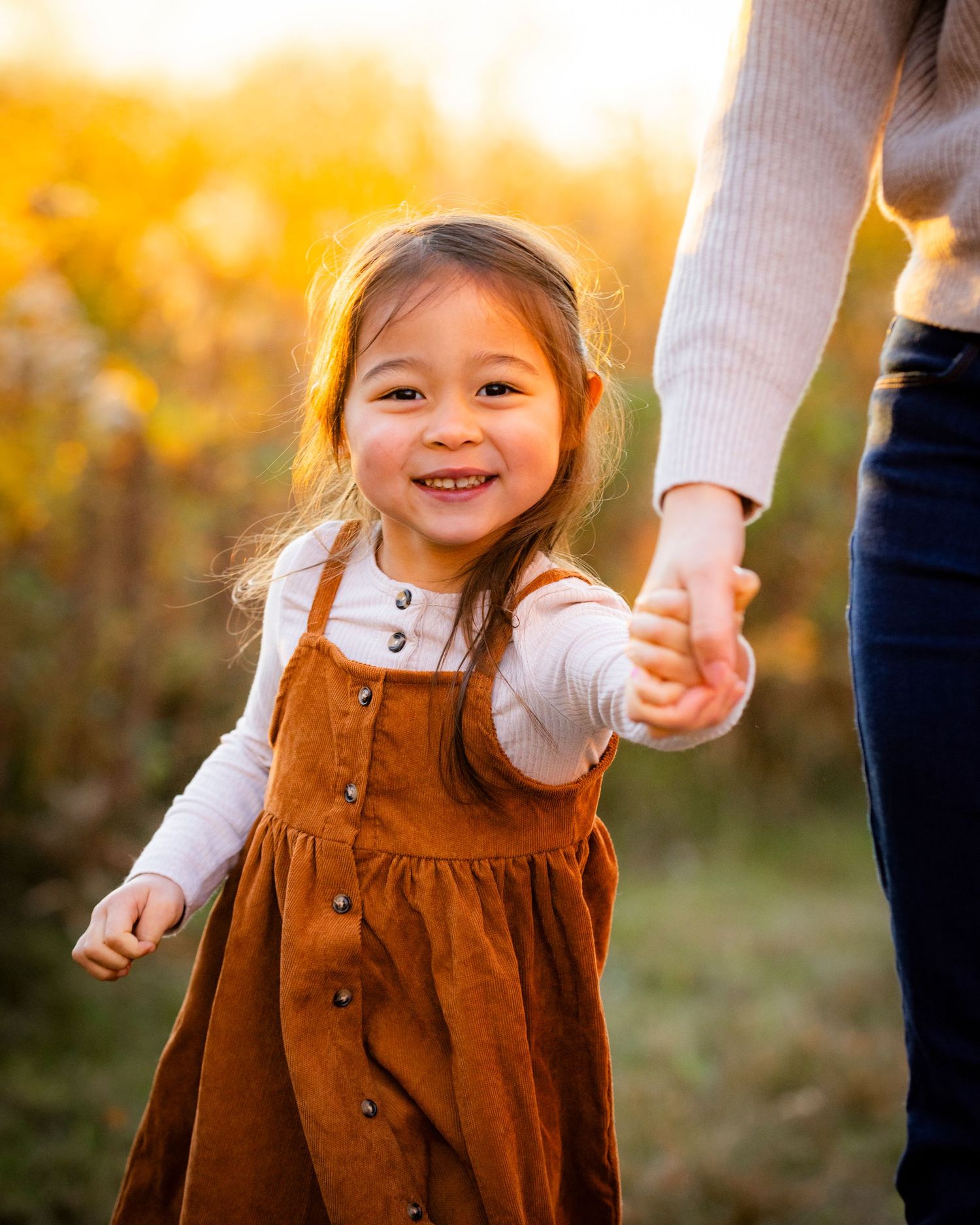 A cheerful little girl in a rust dress holds an adults hand while smiling in golden fall sunlight.
