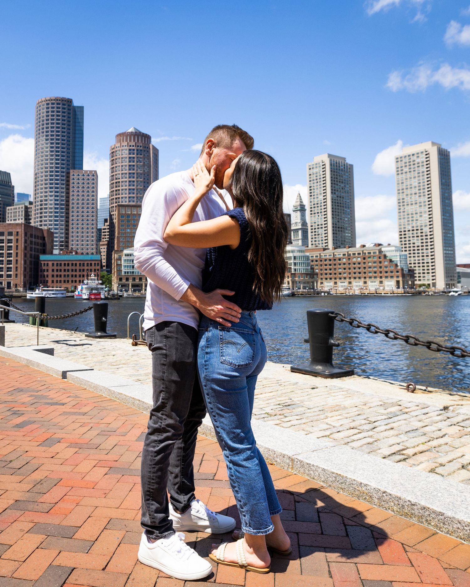 Couple sharing a romantic kiss on Boston harbor waterfront with city skyline in background.