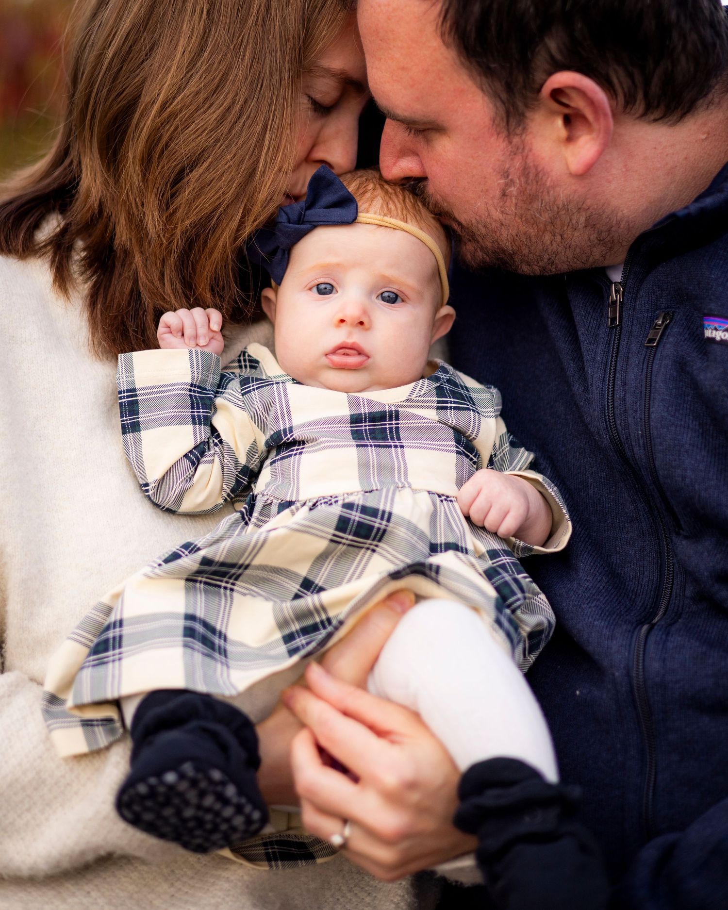 New parents tenderly kiss their infant baby girl in a plaid dress during a fall family portrait.