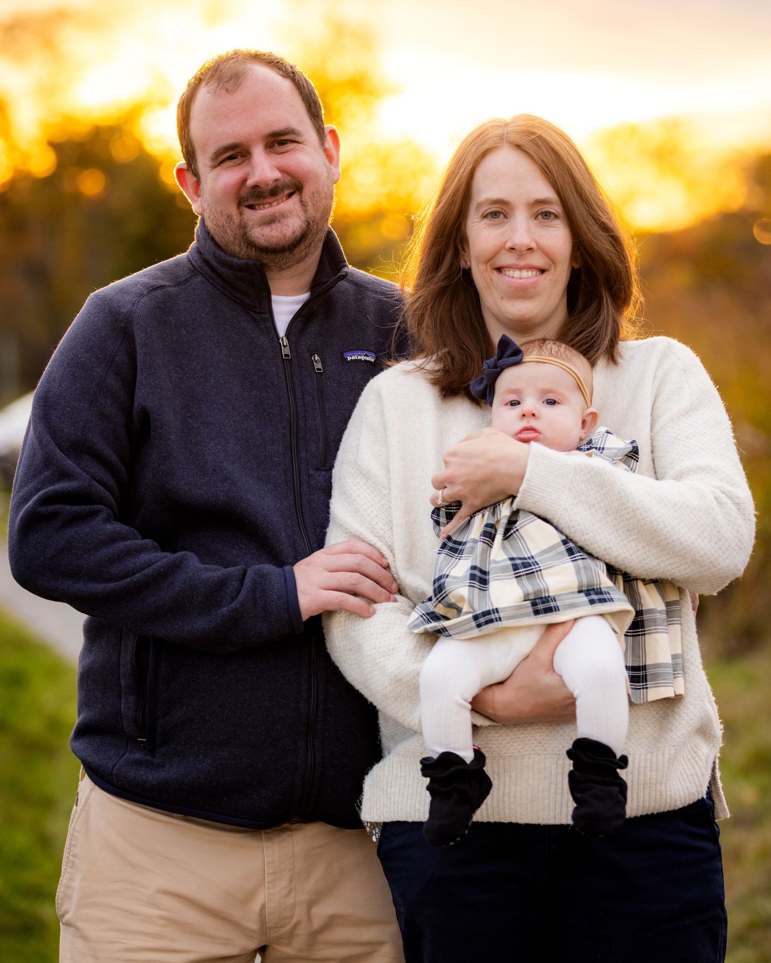 Happy new parents hold their baby girl in a plaid dress during a golden sunset family portrait.