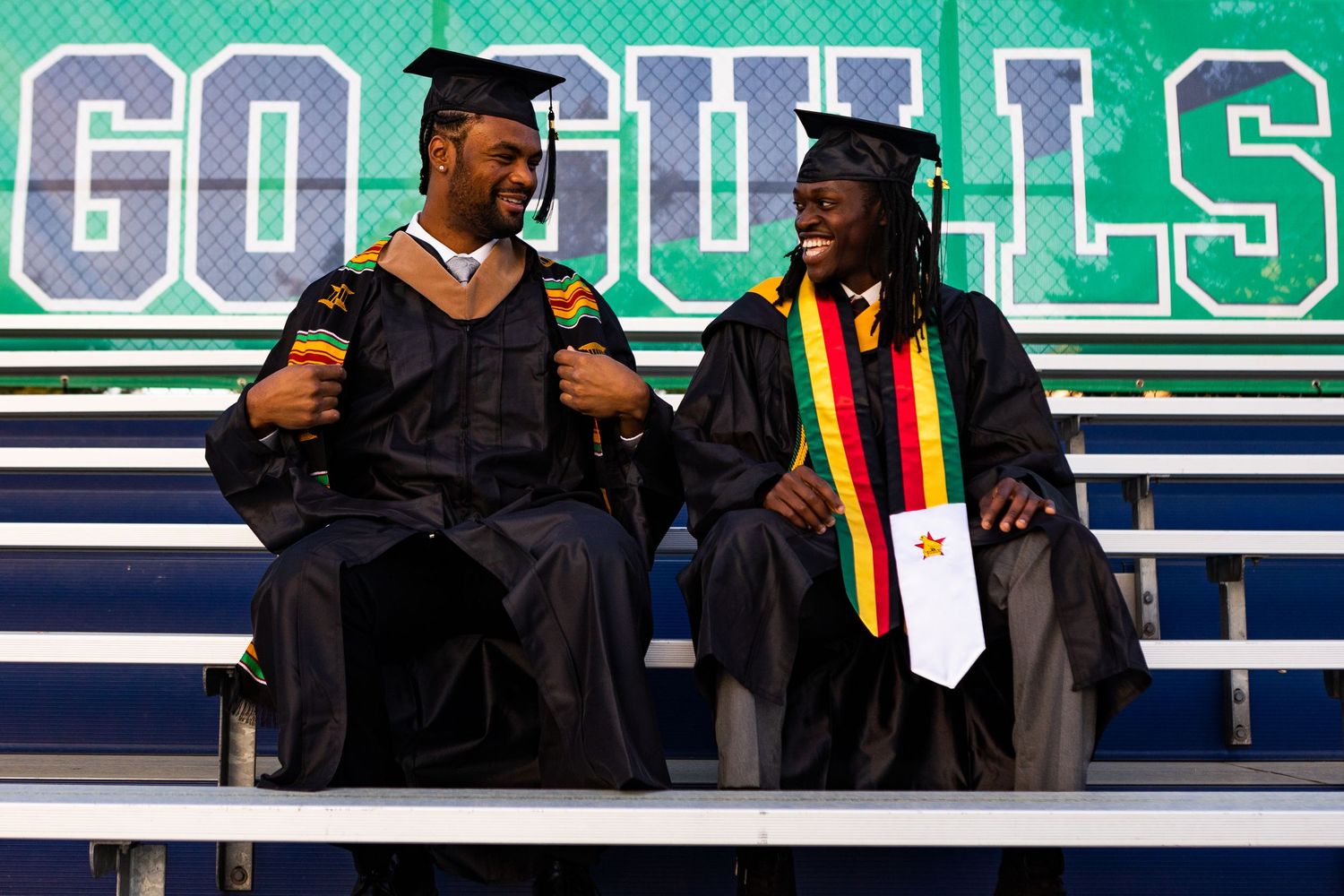 Two male graduates in caps and gowns with Kente stoles laughing together at graduation ceremony.