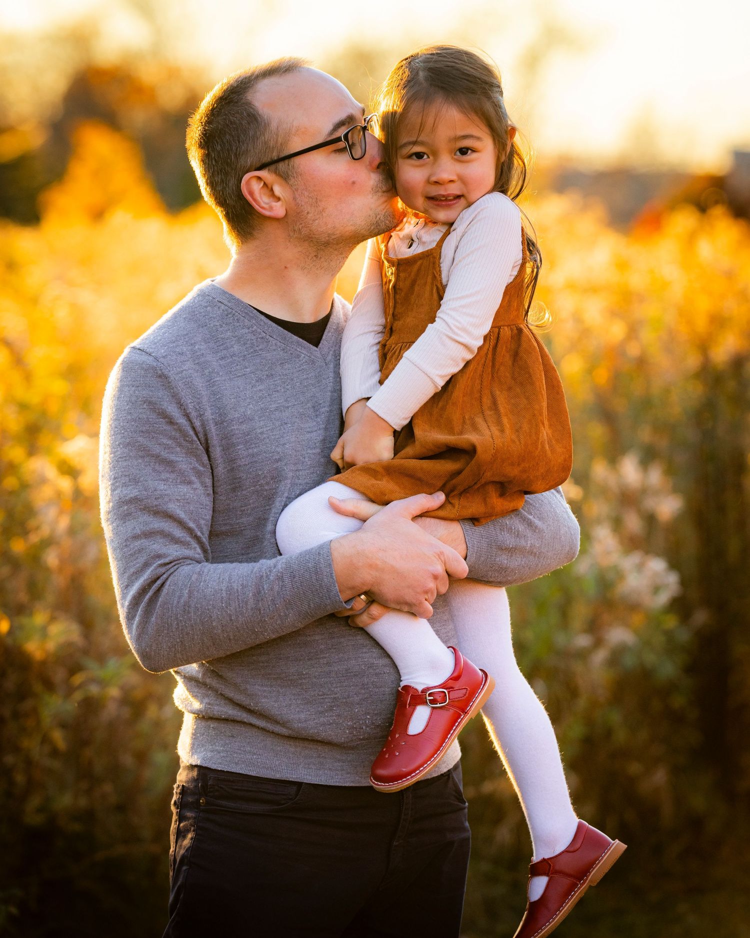 A father tenderly kisses his young daughters cheek while holding her in a sunlit autumn field.