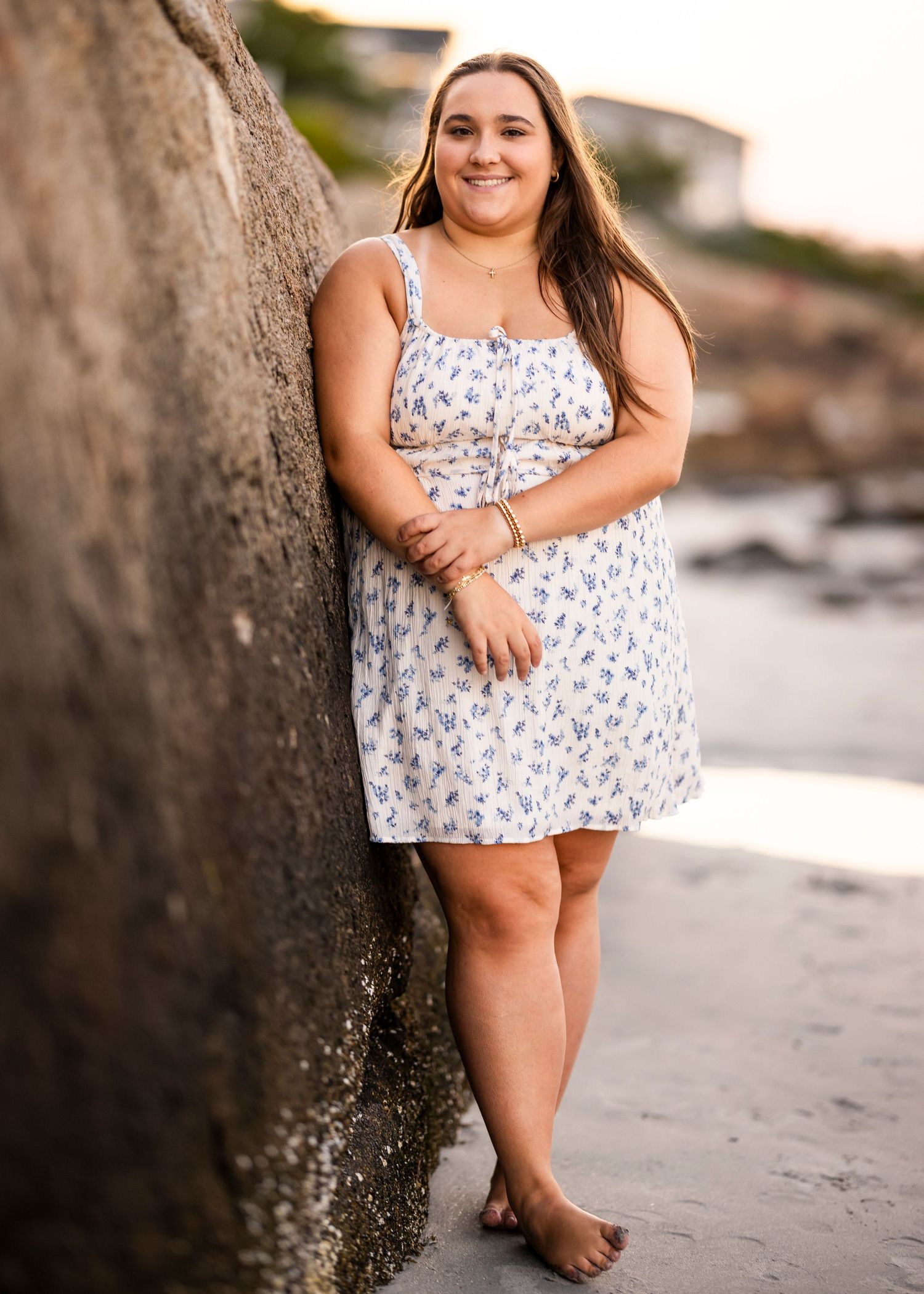 Young woman in a white floral mini dress leans against a large rock during a beach senior portrait session.