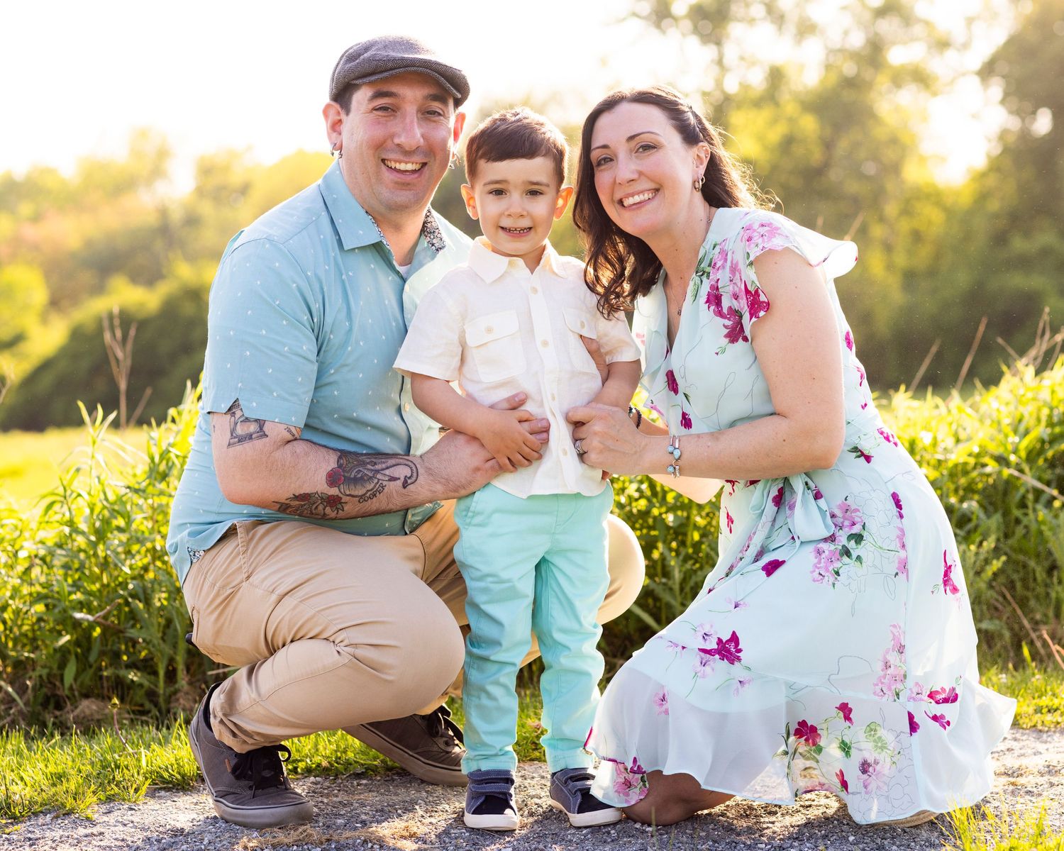 Happy young family poses outdoors in a sunny field, parents kneeling beside their toddler son in a golden hour setting.