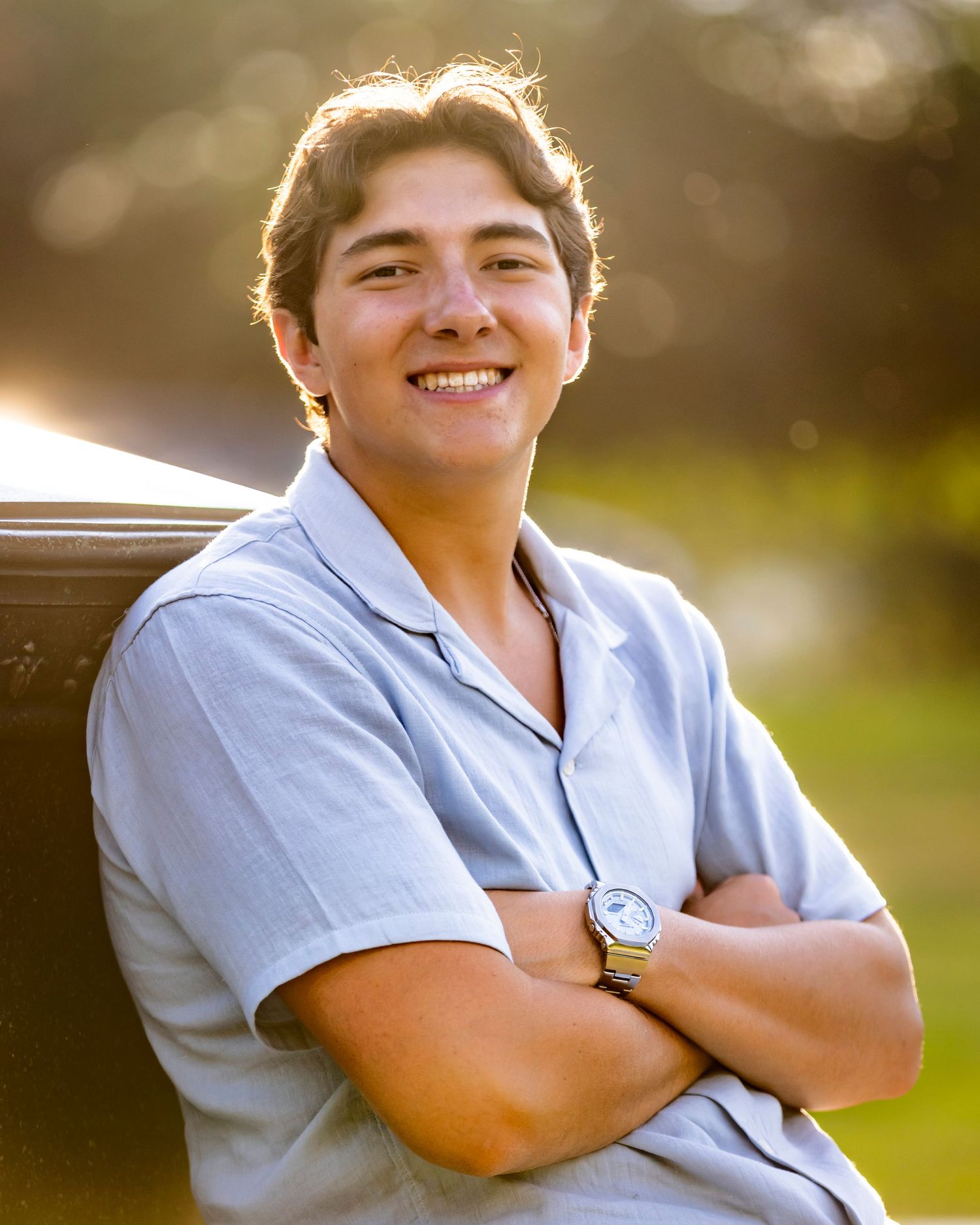 Smiling young man in a light blue shirt with arms crossed leaning against a bench in golden sunlight.
