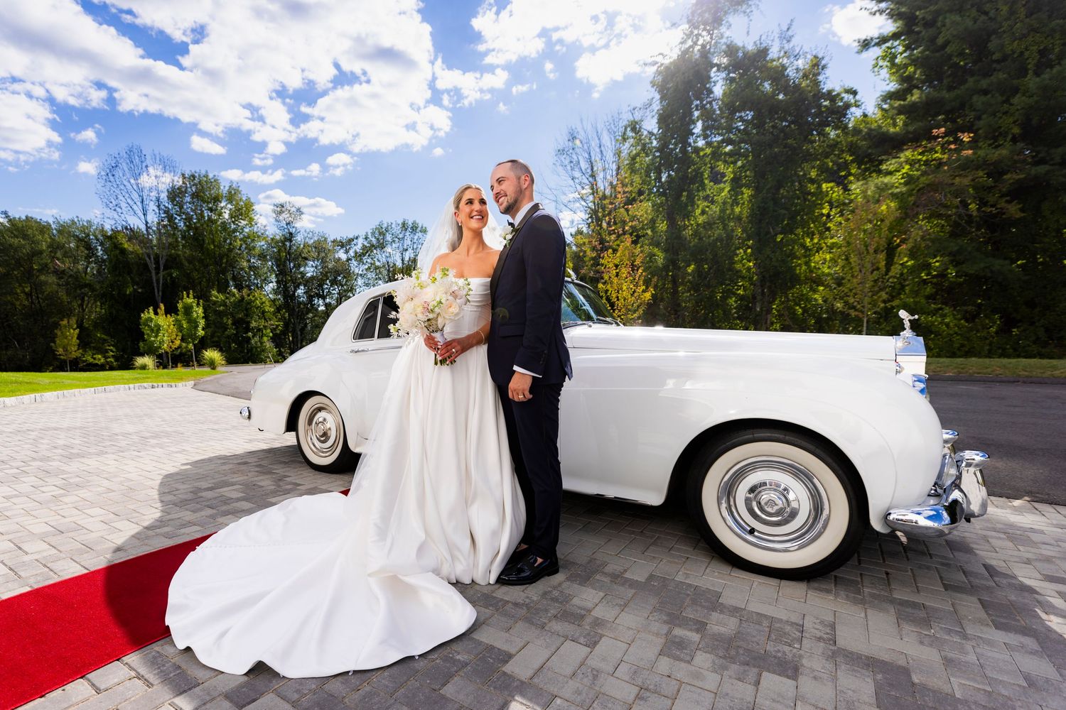 Bride and groom posing beside a classic white Rolls-Royce on a red carpet on their wedding day.