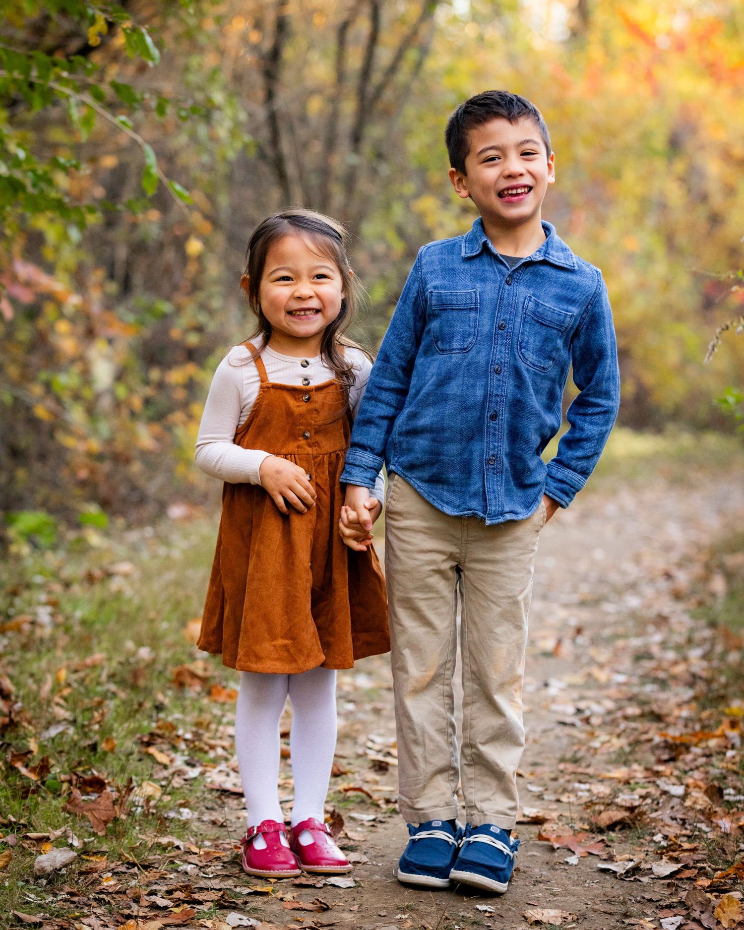 Two young siblings hold hands and smile on a fall nature path surrounded by colorful autumn leaves.