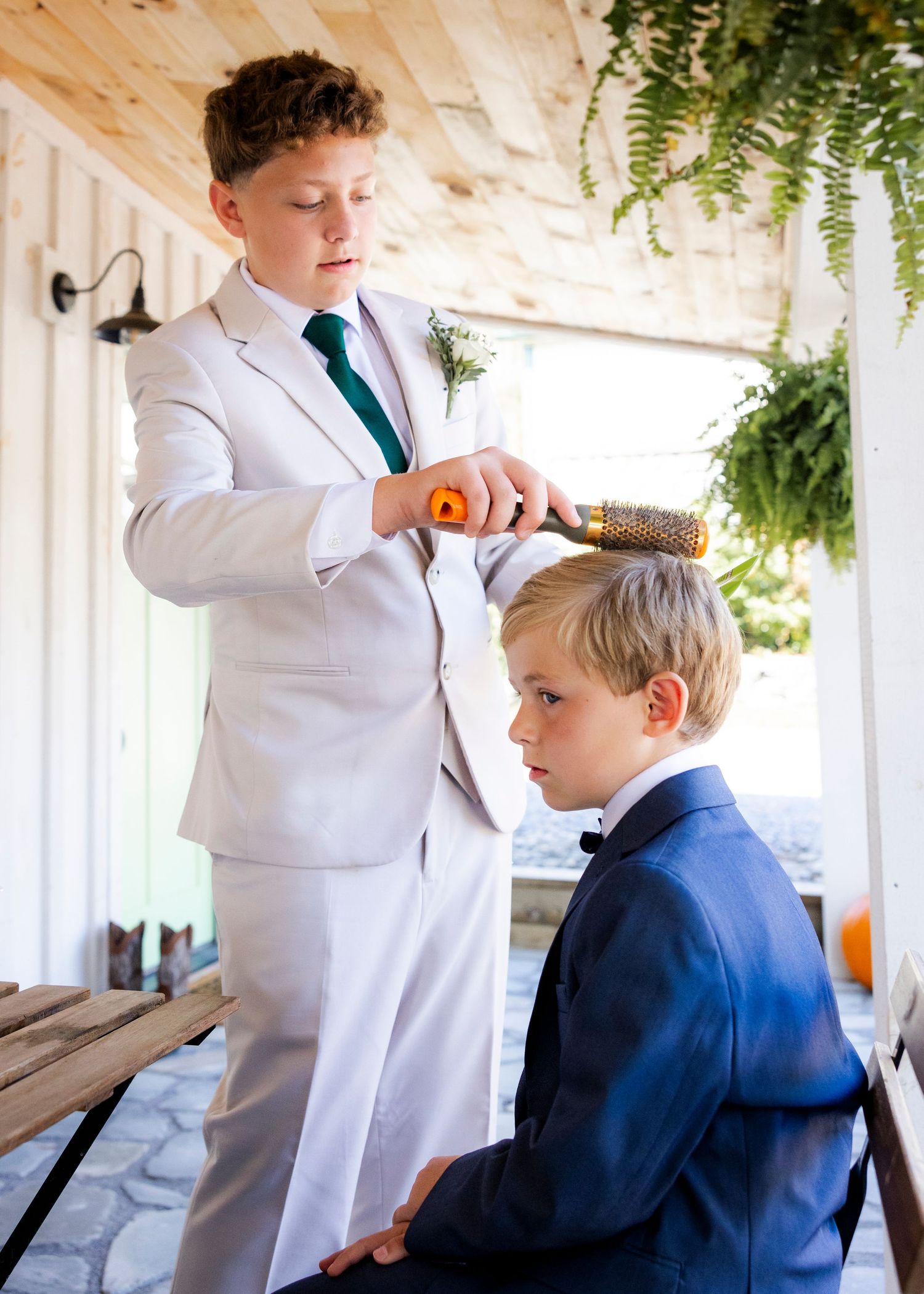 Young boy in white suit brushing hair of younger boy in navy suit at outdoor wedding.