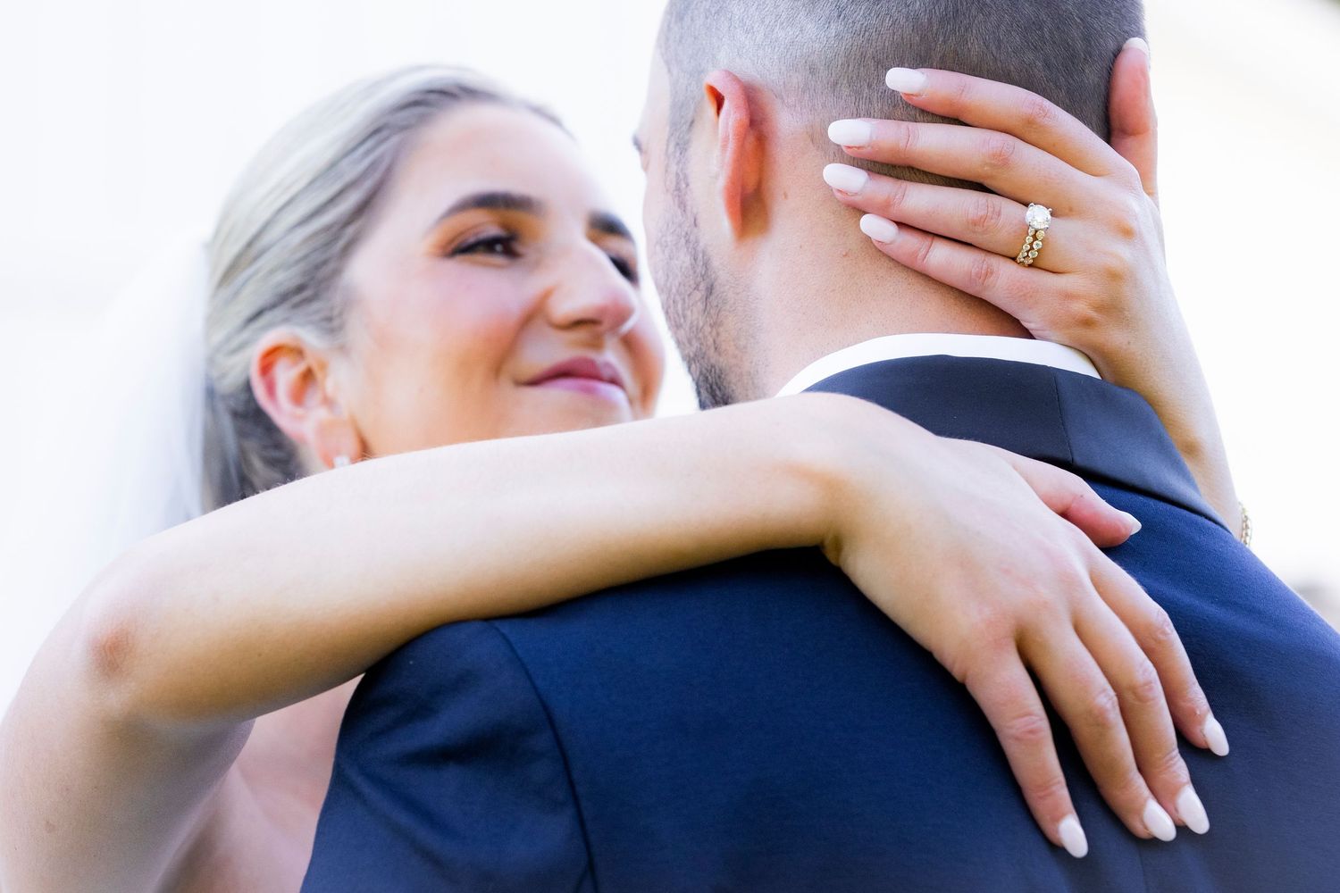 Bride with wedding ring embraces groom tenderly, resting her head on his shoulder outdoors.