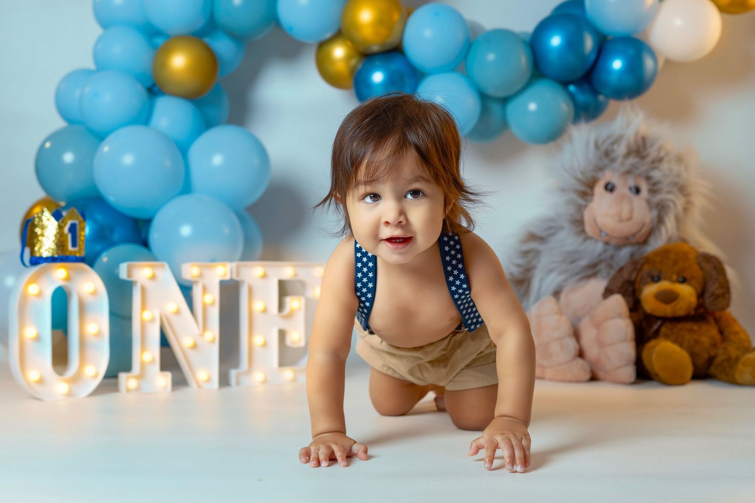 A blue and gold balloon garland decorates a festive cake smash photography setup with white backdrop.