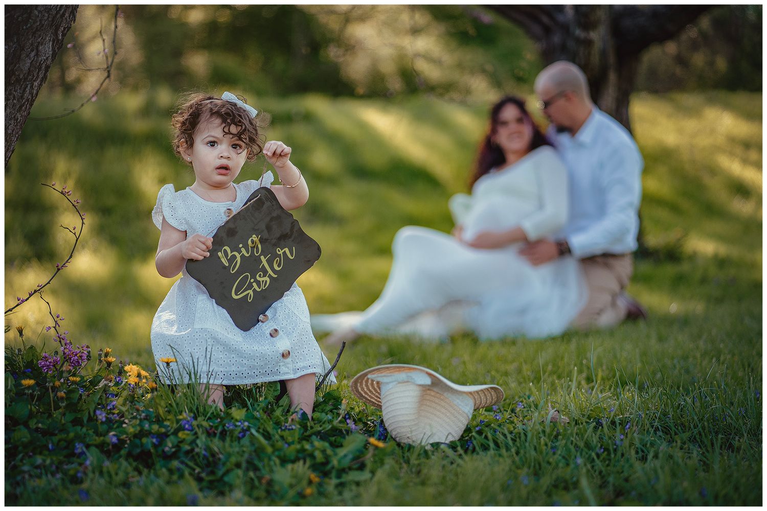 Toddler girl holds Big Sister chalkboard sign while expecting parents embrace softly in green park background.