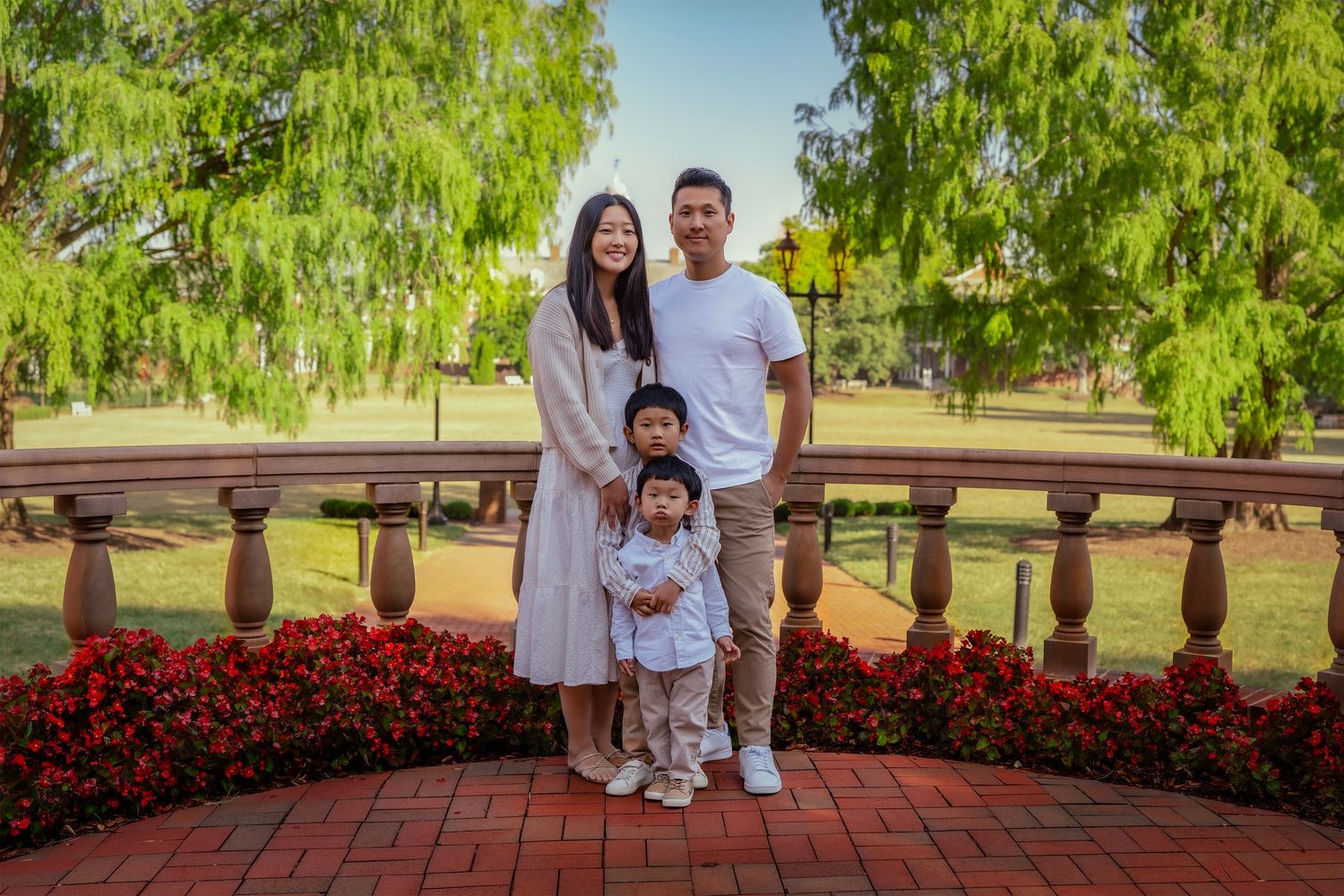 Family of four posing on a brick bridge surrounded by red flowers and green willow trees in a lush park.