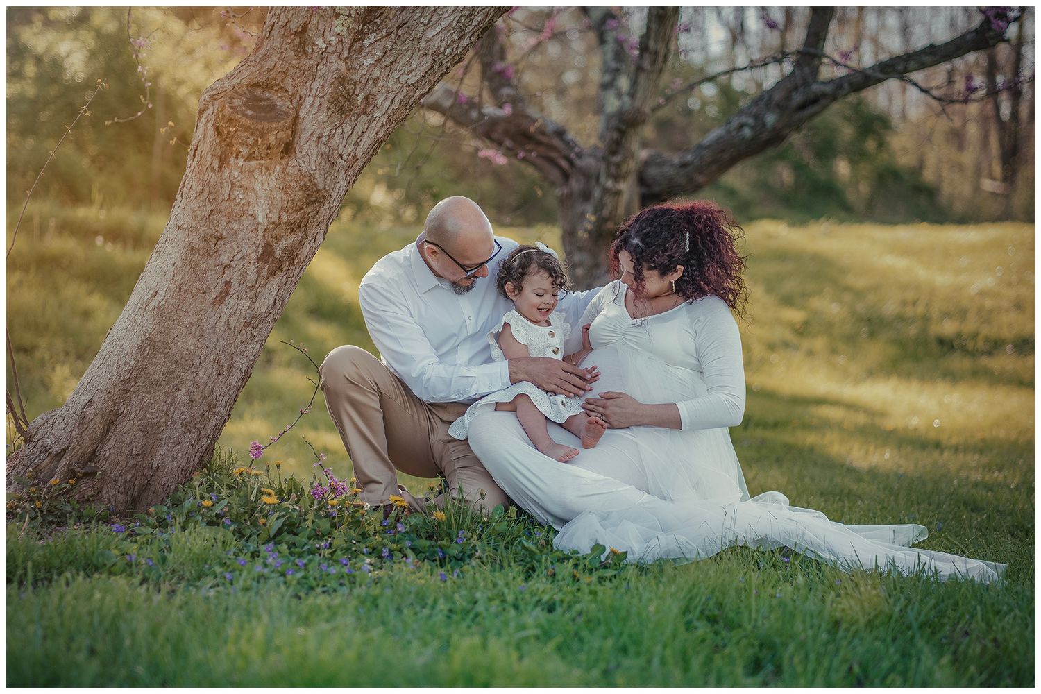 Family of three sitting under a tree in golden hour light, parents in white outfits reading to their toddler on green grass.